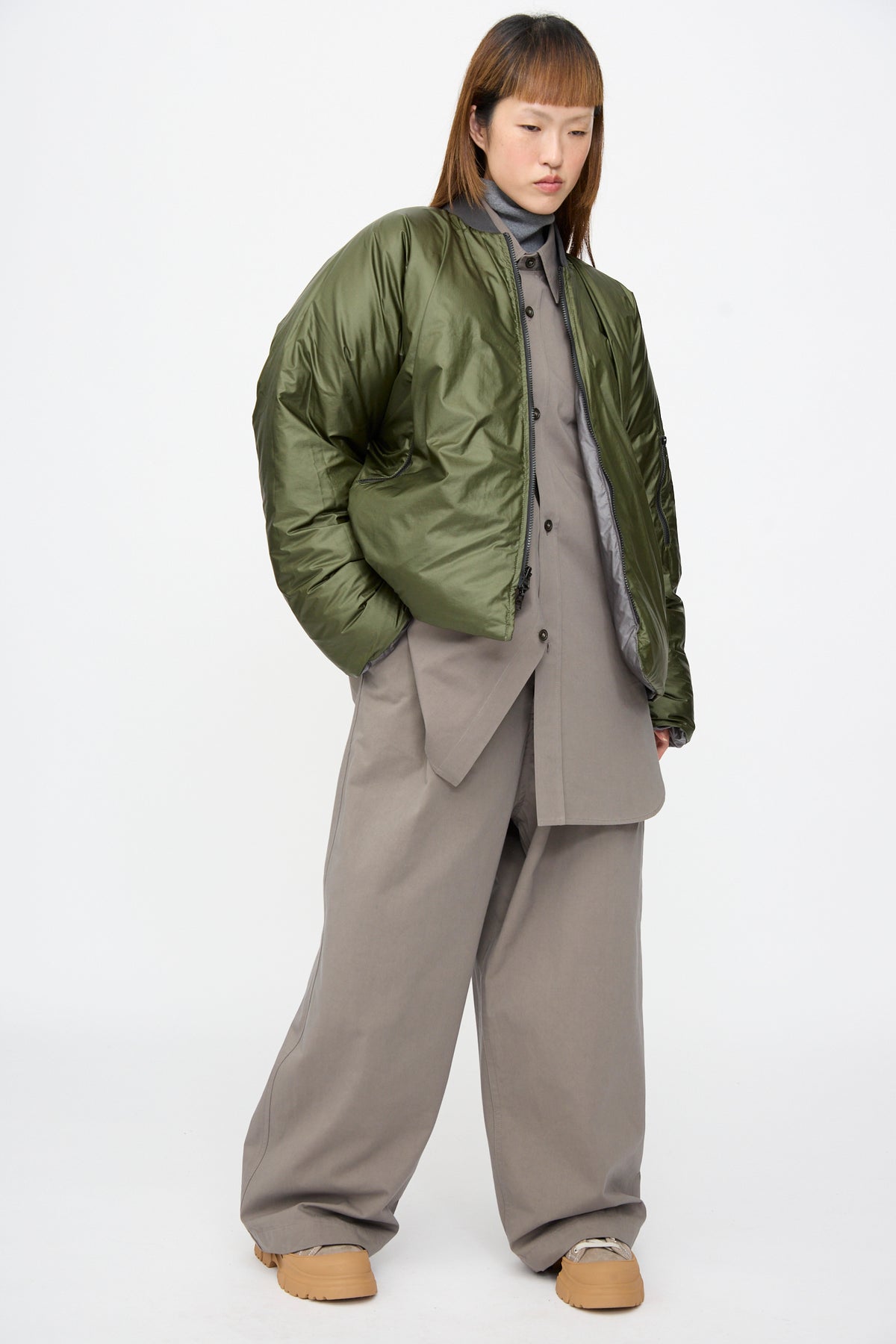 A person wears an olive green, reversible Sofie D’Hoore Origin Down Jacket in Charcoal and Khaki over a grey collared shirt, wide-leg trousers, and tan platform shoes.