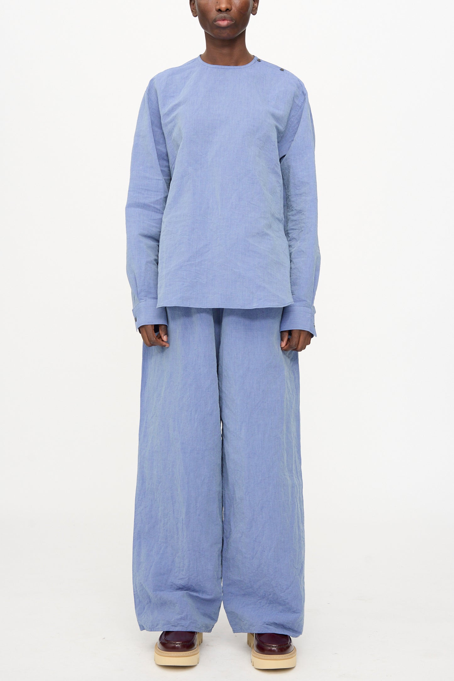 A person wears the Sofie D'Hoore Pavon Pant in Blue Jean with a loose light blue top and burgundy platform shoes, standing against a plain white background.