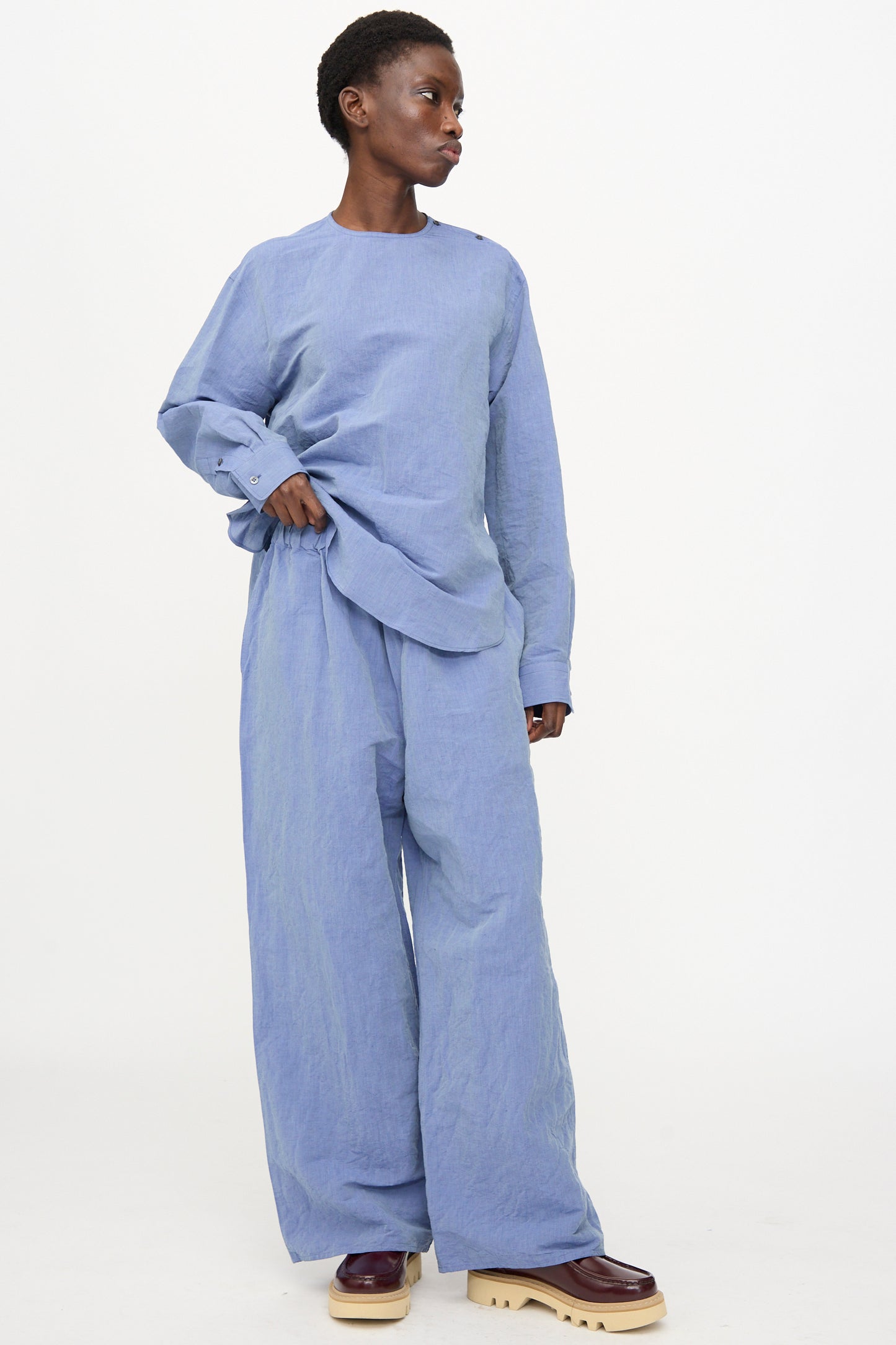 A person stands against a white background wearing Sofie D'Hoore's Pavon Pant in Blue Jean, a loose-fitting, oversized linen pant with an elastic waist, paired with a light blue long-sleeve top and burgundy platform shoes.