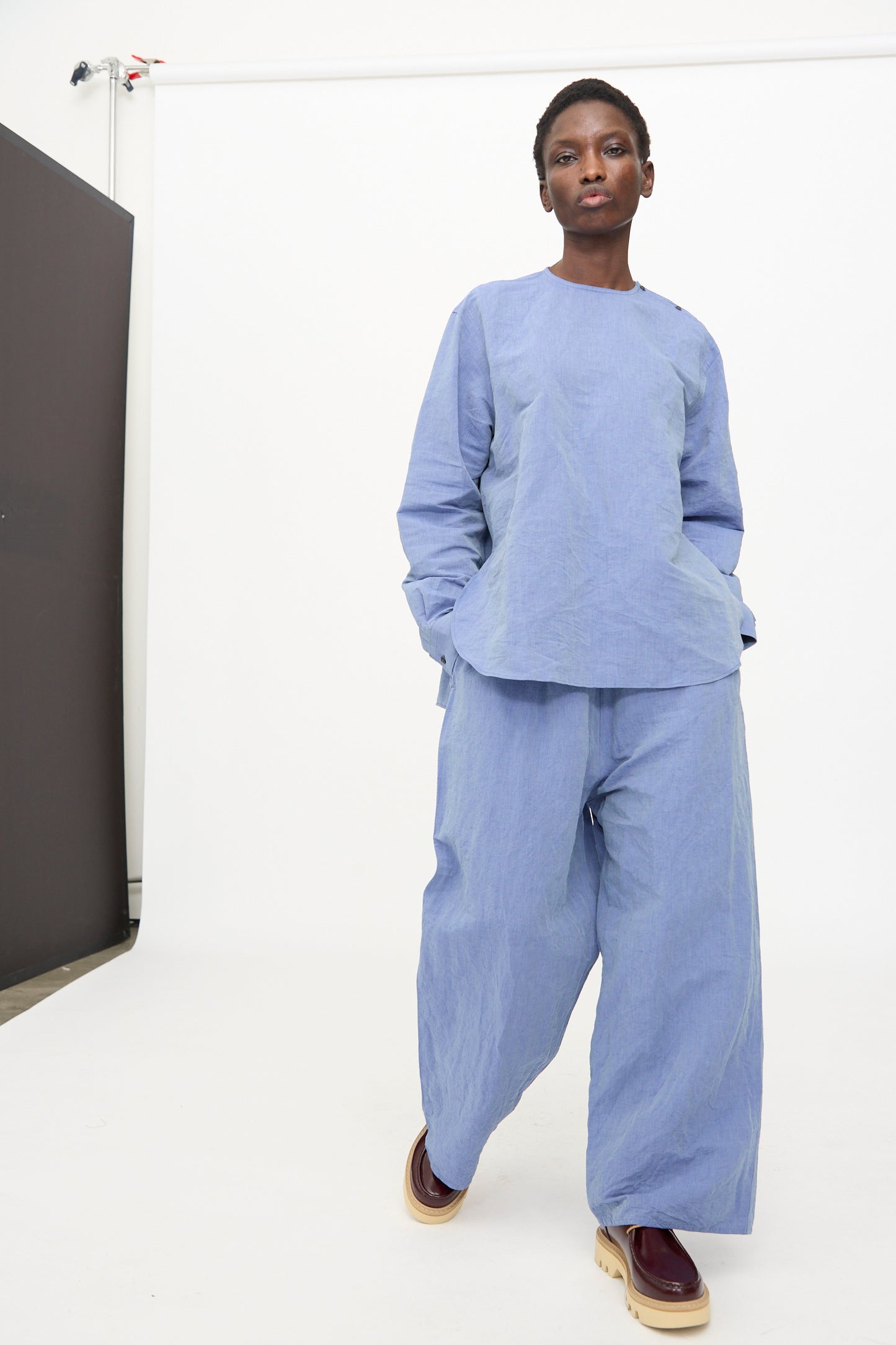A person stands before a white backdrop wearing the Sofie D'Hoore Pavon Pant in Blue Jean with a loose light blue top and brown platform shoes.