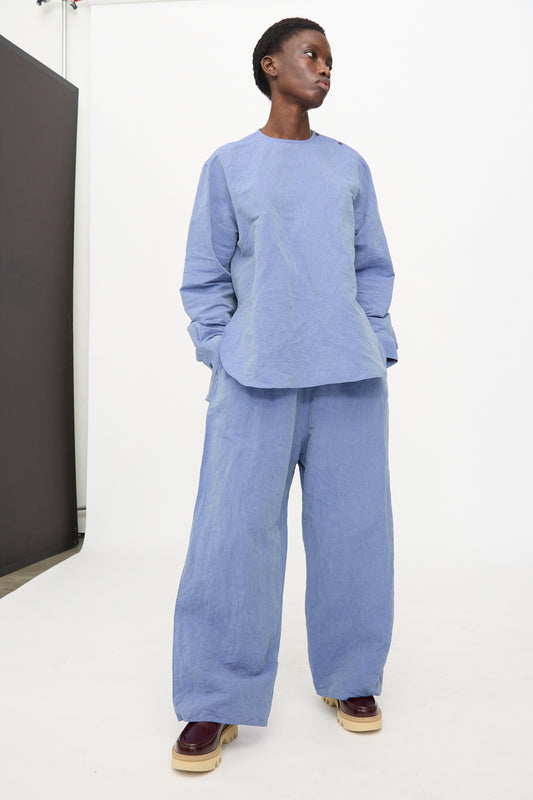Model wears the Sofie D'Hoore Pavon Pant in Blue Jean—oversized, wide-leg linen pants—paired with a loose-fitting, long-sleeve light blue top. She stands against a white background with hands in her pockets.