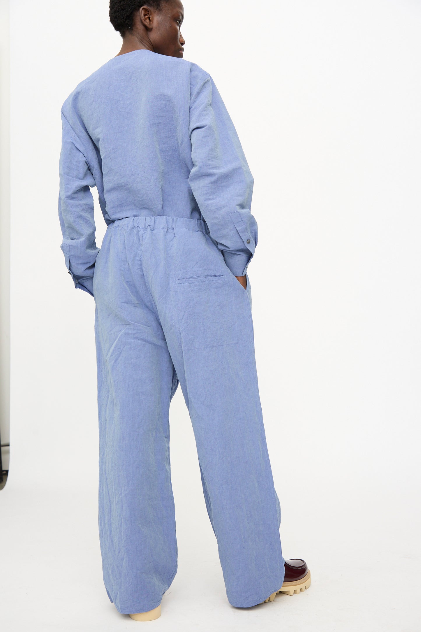 A person stands with hands in pockets, wearing the Sofie D'Hoore Pavon Pant in Blue Jean and cream-soled shoes, facing away against a plain white background.