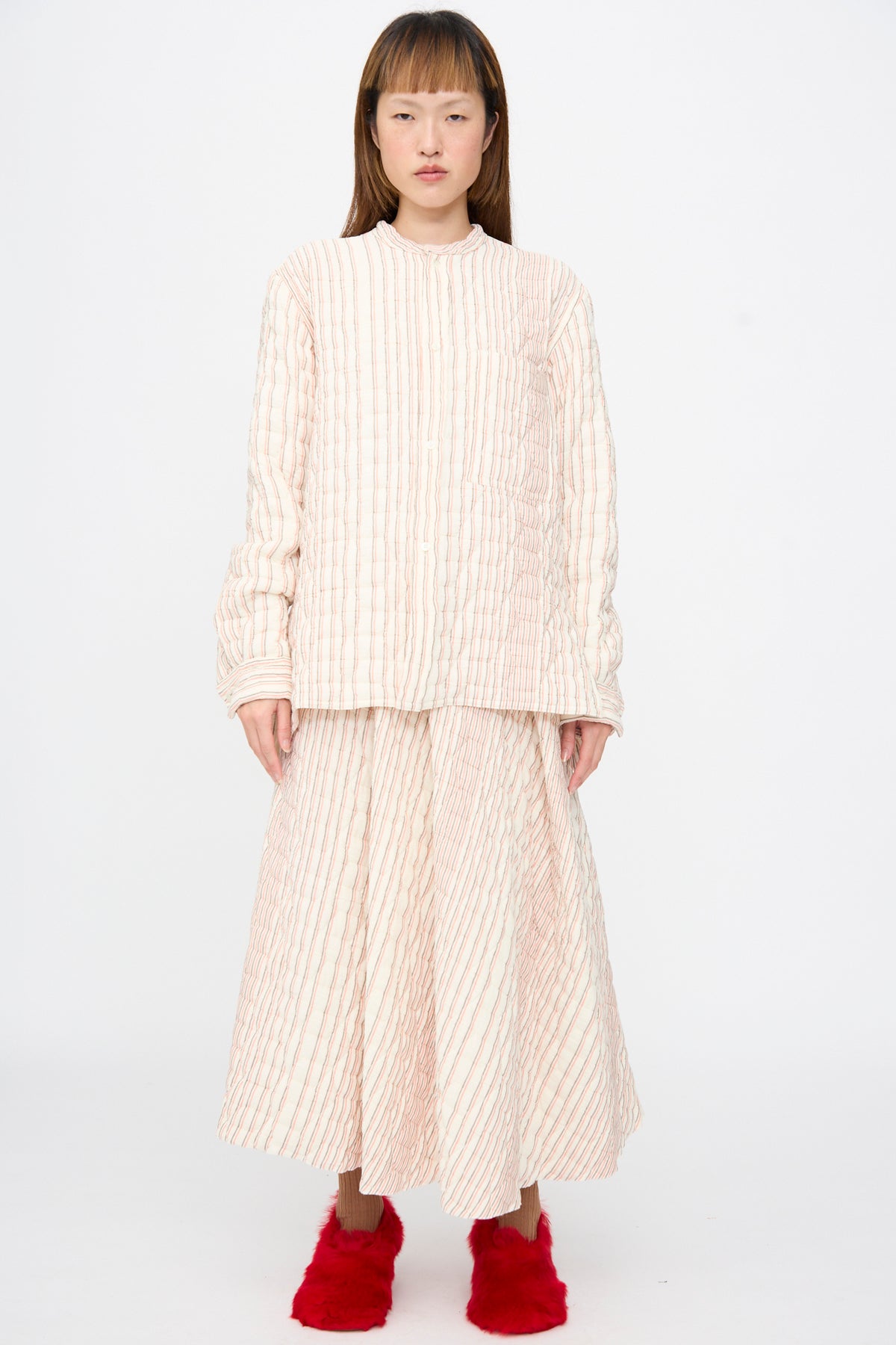 A person wearing a cream and red striped Sofie D’Hoore Quilted Cotton Bela Shirt with a matching pleated skirt and bright red fuzzy shoes.