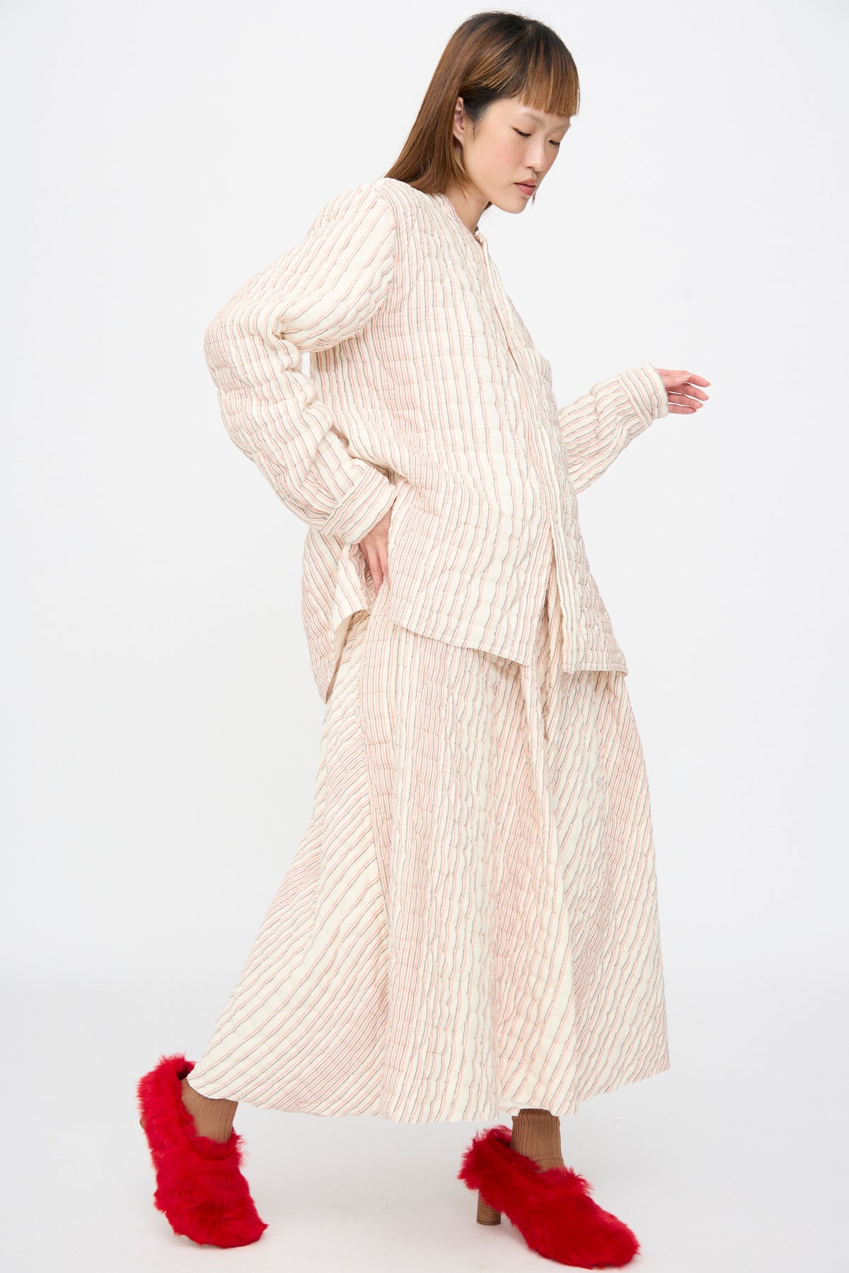 A person wearing a cream quilted cotton top and a long Quilted Cotton Bela Shirt in Red Stripe, paired with bright red faux-fur mules.