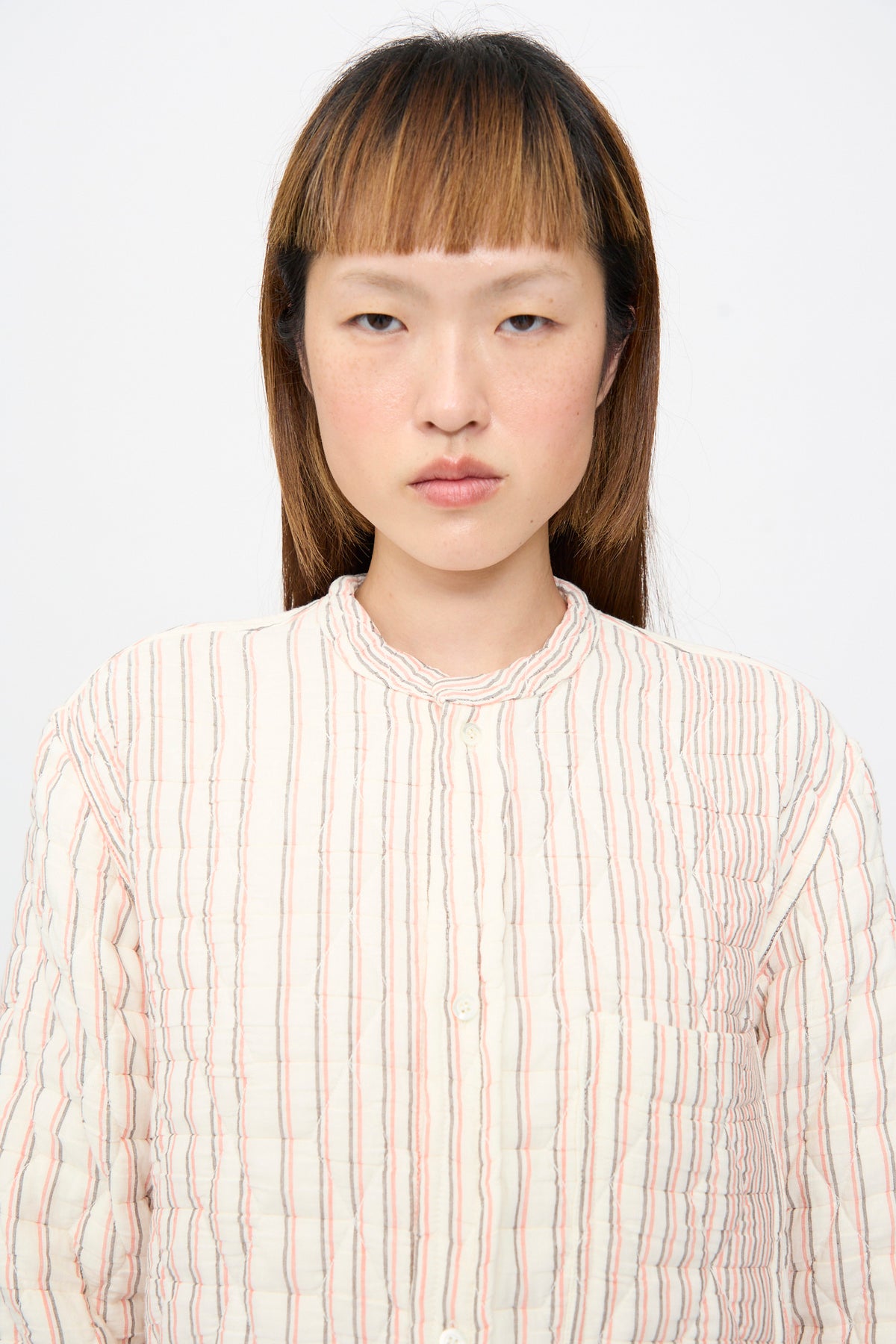 A person wearing a Sofie D’Hoore Quilted Cotton Bela Shirt in Red Stripe, a relaxed fit, cream-colored quilted cotton top with thin red and grey vertical stripes and a band collar.