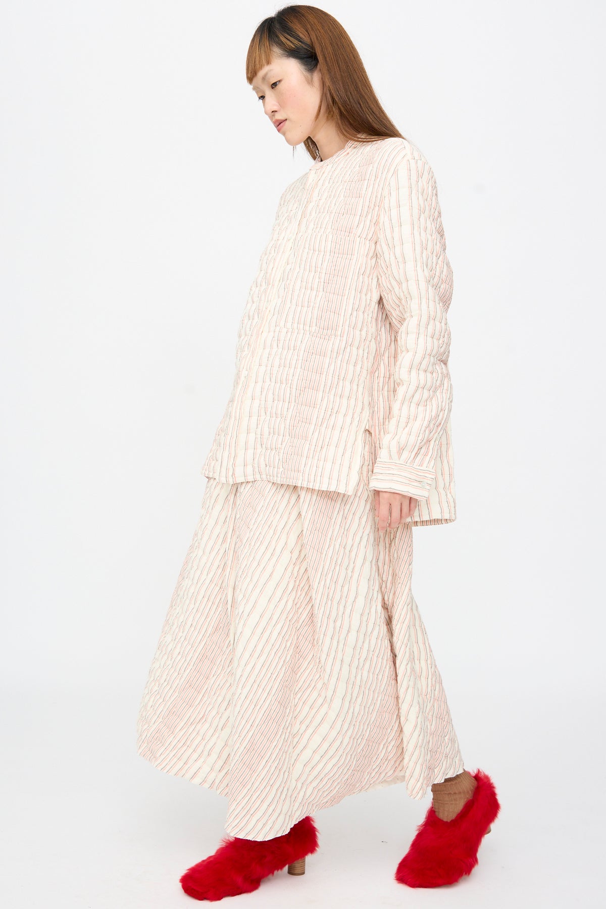 A person wears a quilted, long-sleeved cream and red vertical-striped top with a matching Quilted Cotton Savy Skirt in Red Stripe and fuzzy red slip-on shoes.