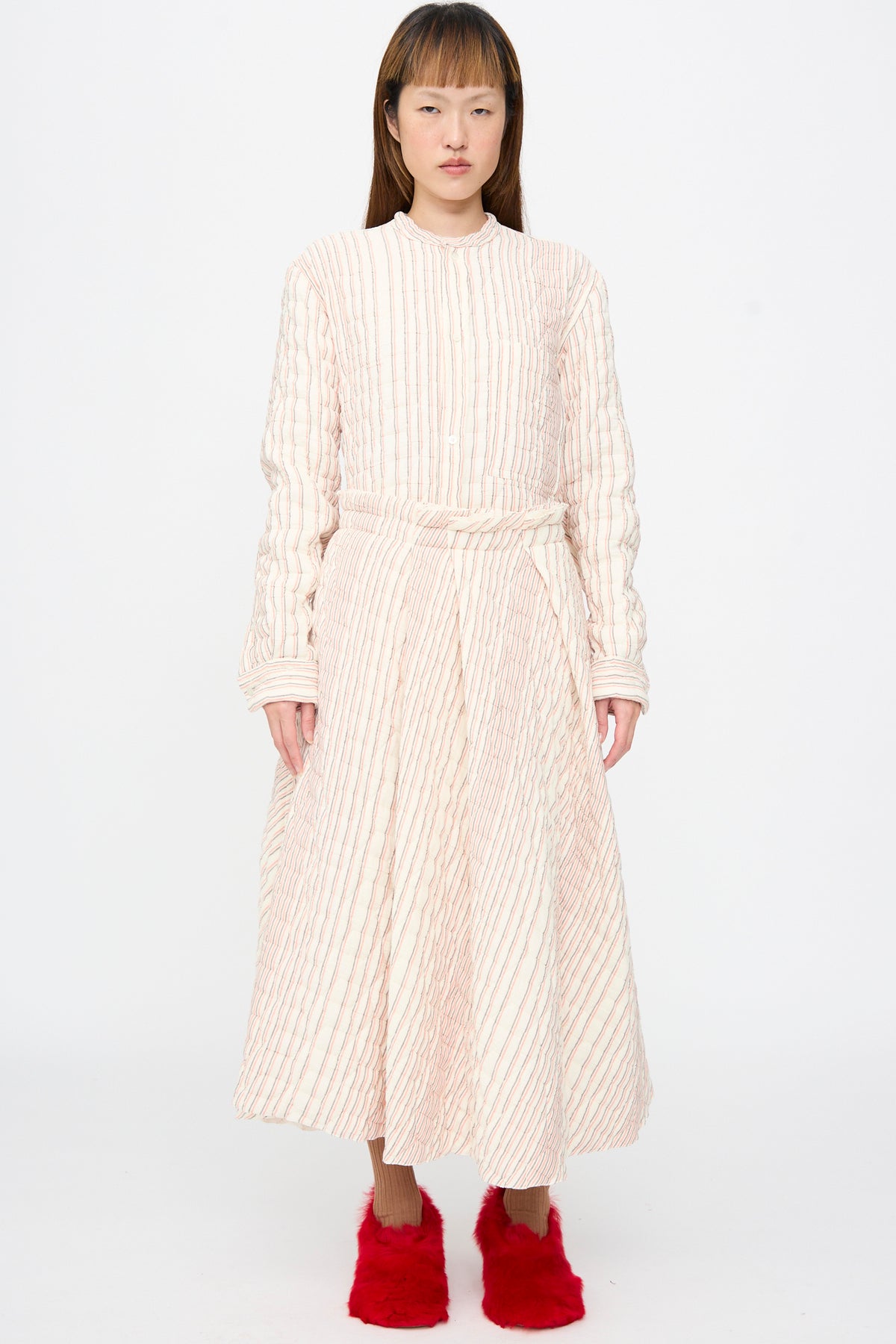 A person wears a Sofie D’Hoore Quilted Cotton Savy Skirt in Red Stripe, a long-sleeved, quilted A-line midi set with a thin red striped pattern and bright red fuzzy shoes.