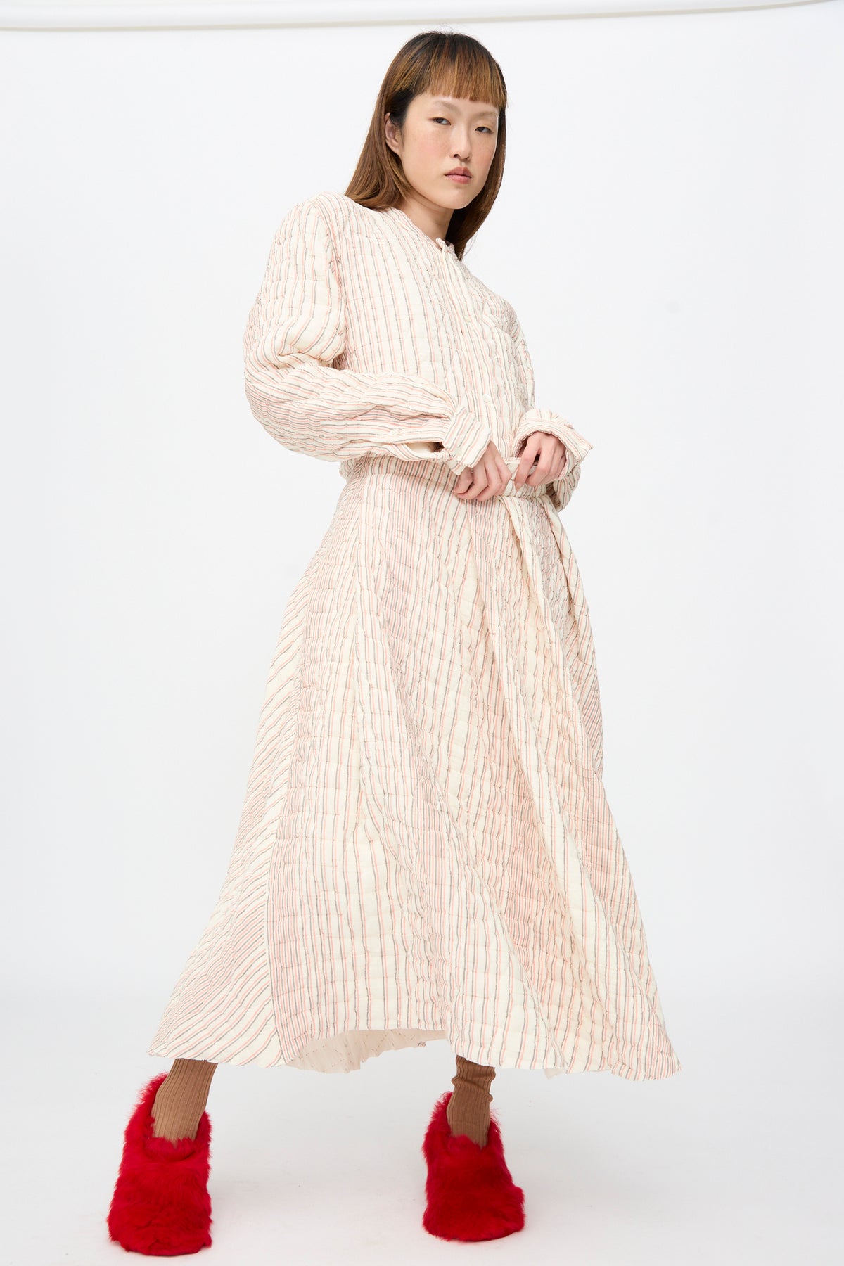 A model wears a Sofie D’Hoore Quilted Cotton Savy Skirt in Red Stripe, a long-sleeved A-line maxi skirt with thin red stripes, paired with bright red fuzzy slippers.