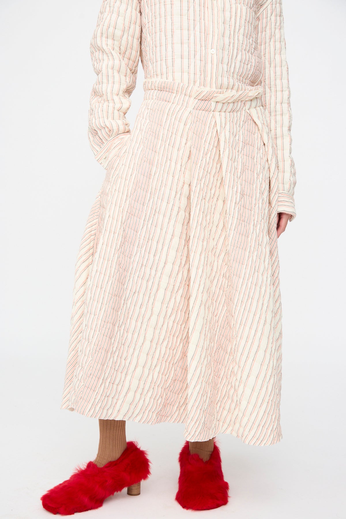 A person wears a Sofie D’Hoore Quilted Cotton Savy Skirt in Red Stripe and a matching shirt with thin vertical red stripes, paired with brown ribbed socks and bright red faux-fur mules.