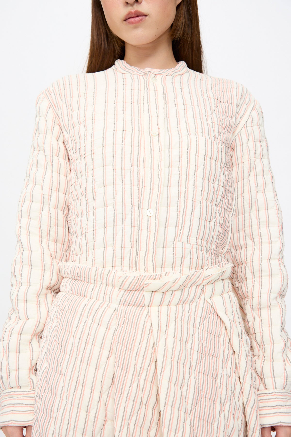 A person wearing a Quilted Cotton Savy Skirt in Red Stripe, styled as a long-sleeved, cream-colored quilted shirt with thin red and grey vertical stripes, an A-line silhouette, and a belted waist.