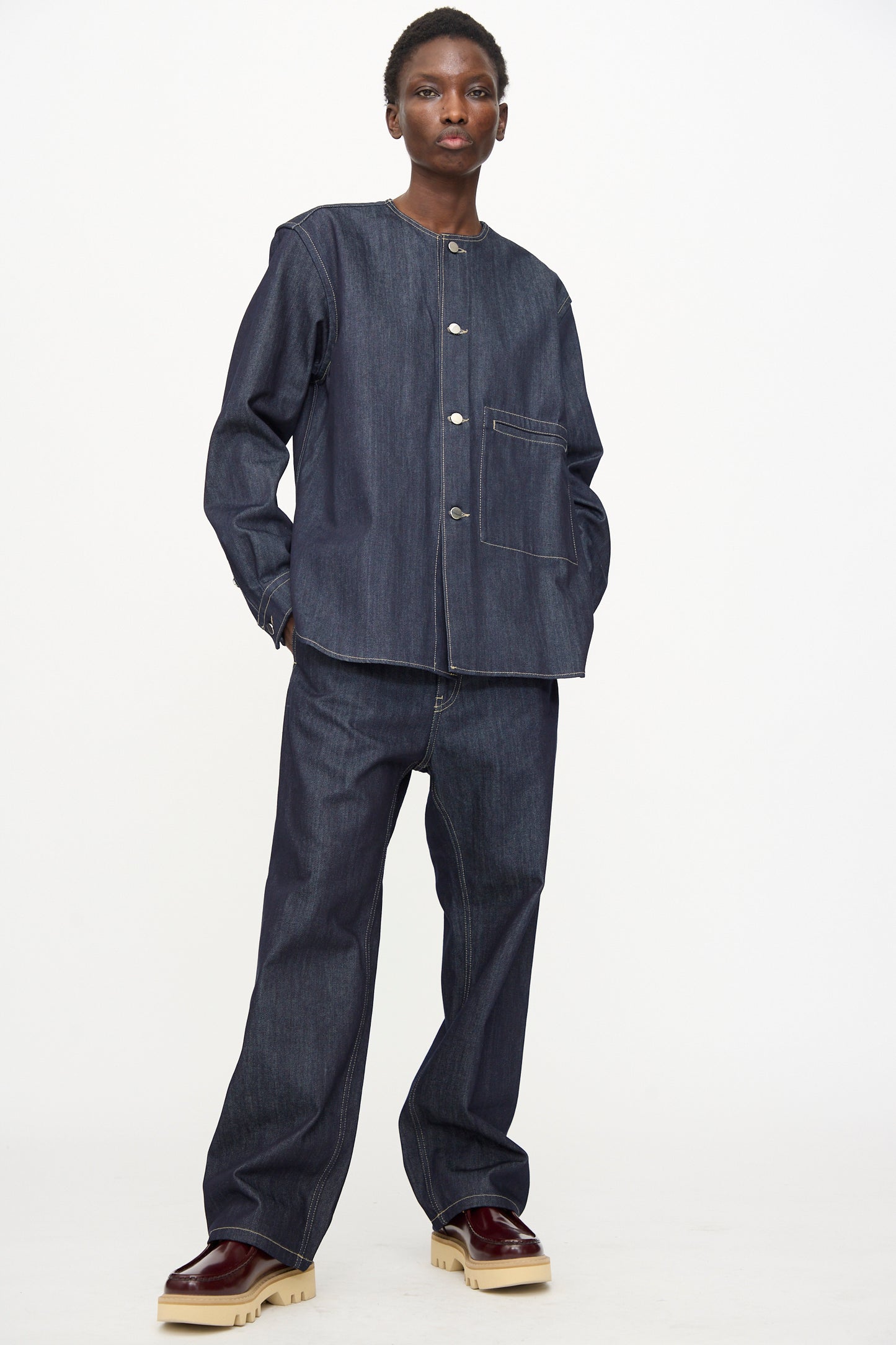 A person stands against a plain background in Sofie D’Hoore’s Raw Denim Pasay Pant in Midnight—straight leg, raw cotton denim—paired with a relaxed fit dark denim jacket and maroon shoes with thick, light soles.