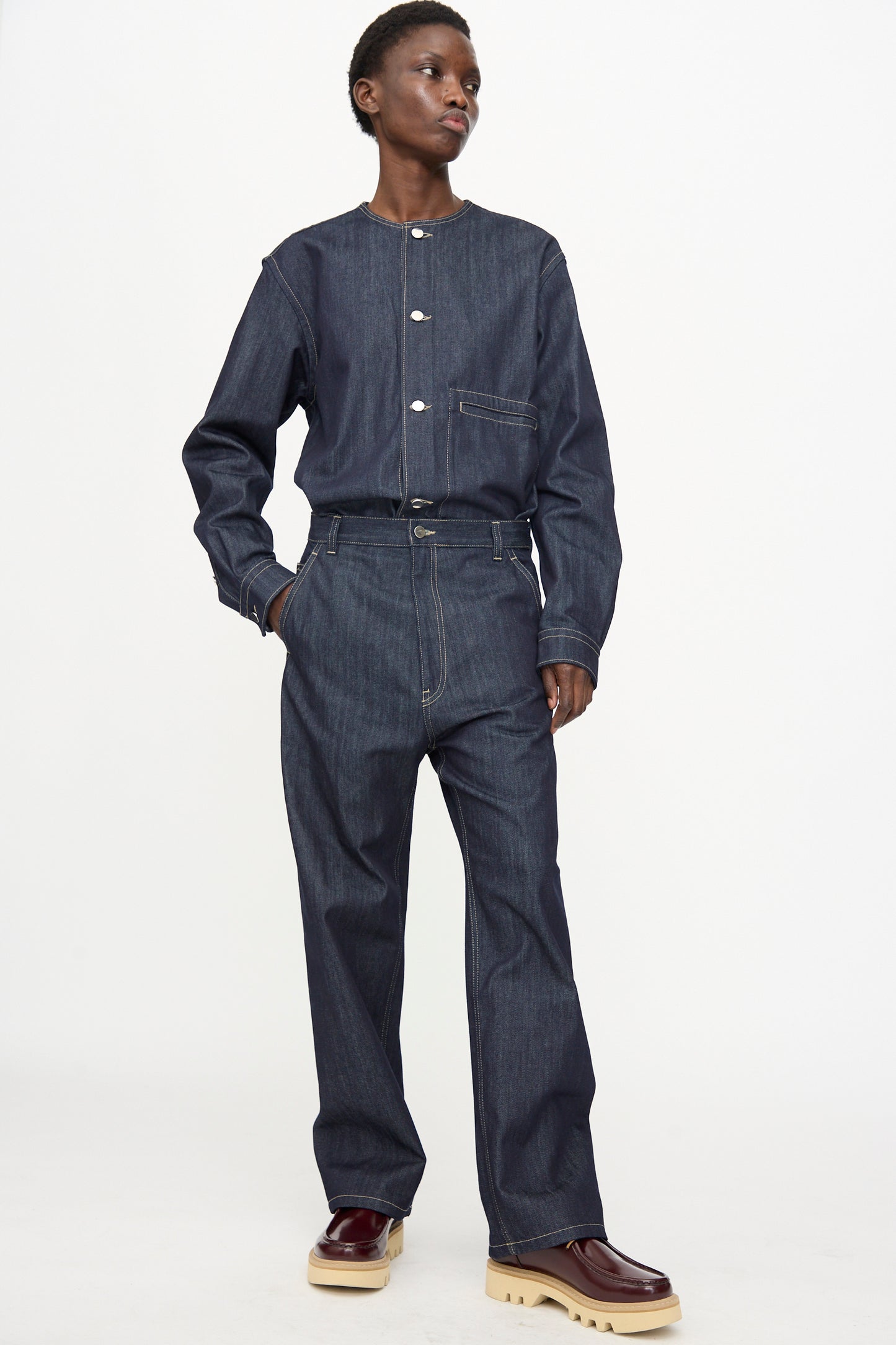 A person wears Sofie D'Hoore's Raw Denim Pasay Pant in Midnight, styled with a matching dark raw cotton denim shirt and burgundy shoes with chunky beige soles, standing against a plain background.