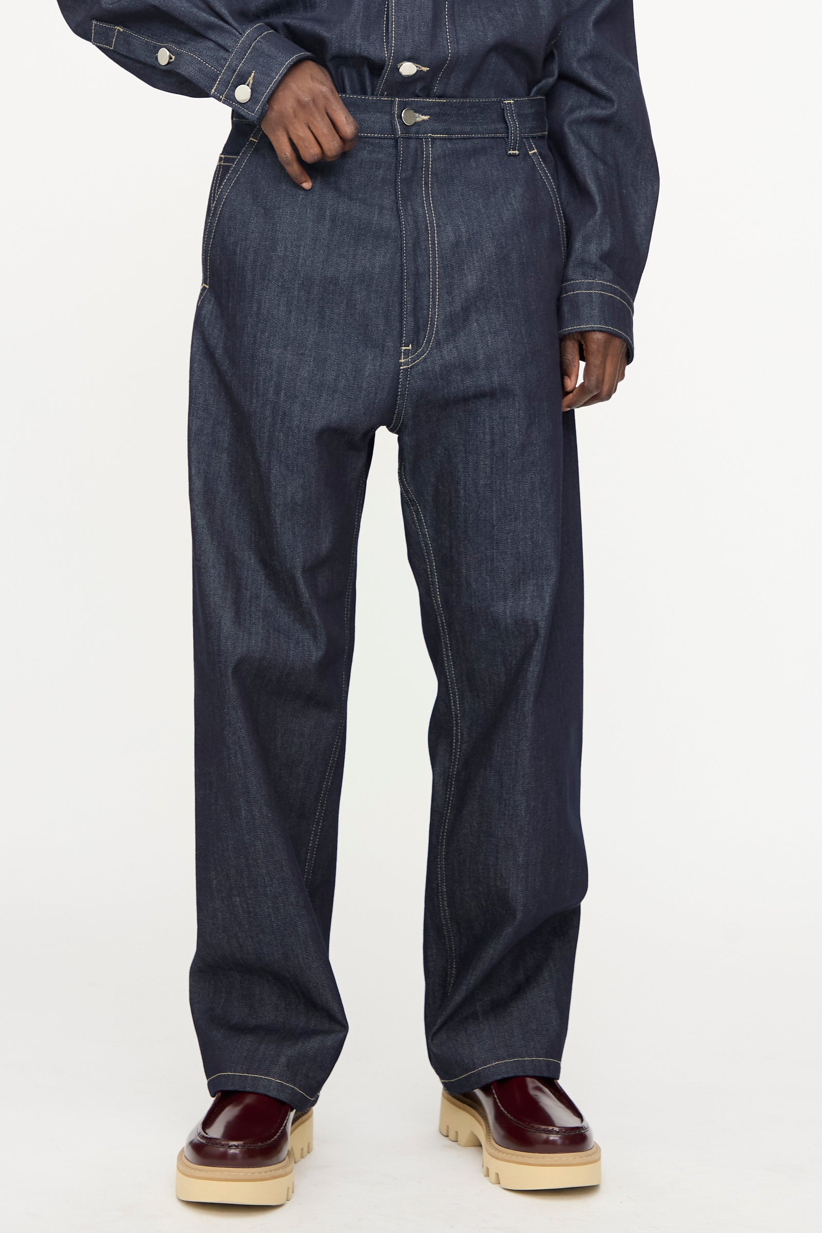 Wearing Sofie D'Hoore Raw Denim Pasay Pant in Midnight—dark blue, relaxed fit raw cotton jeans with white stitching—paired with a matching denim shirt and maroon chunky shoes.