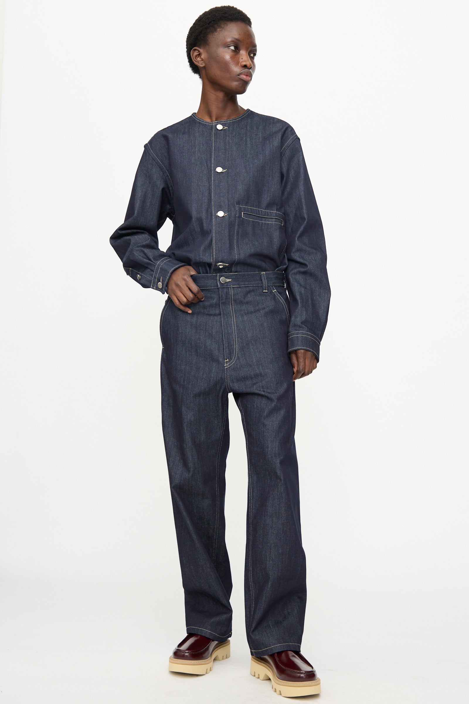 A person wearing Sofie D'Hoore's Raw Denim Pasay Pant in Midnight styled with a relaxed fit dark denim shirt and burgundy platform shoes, standing against a plain white background.