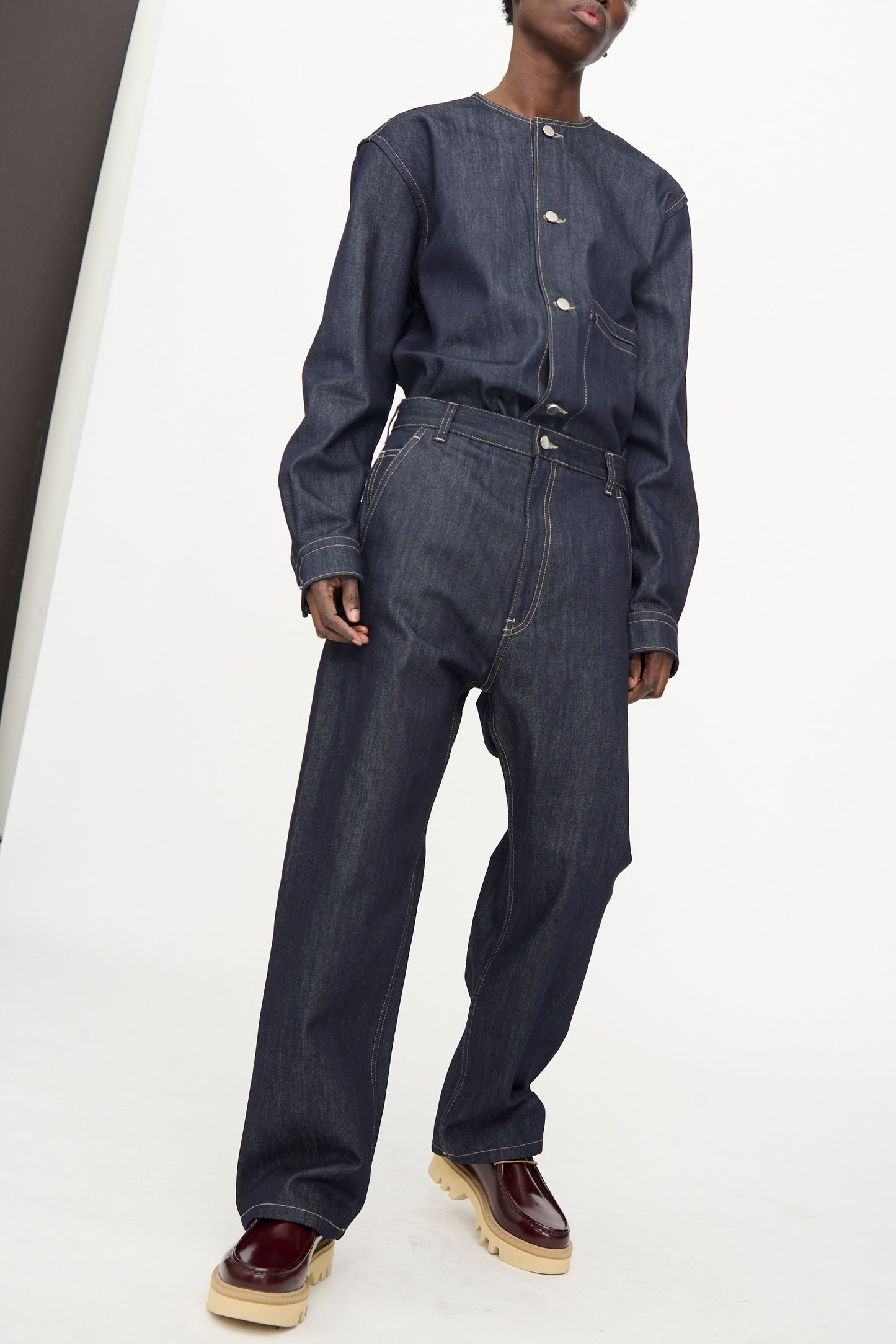 A person stands against a plain white background wearing Sofie D’Hoore’s Raw Denim Pasay Pant in Midnight with a matching dark raw denim jacket and burgundy platform shoes.
