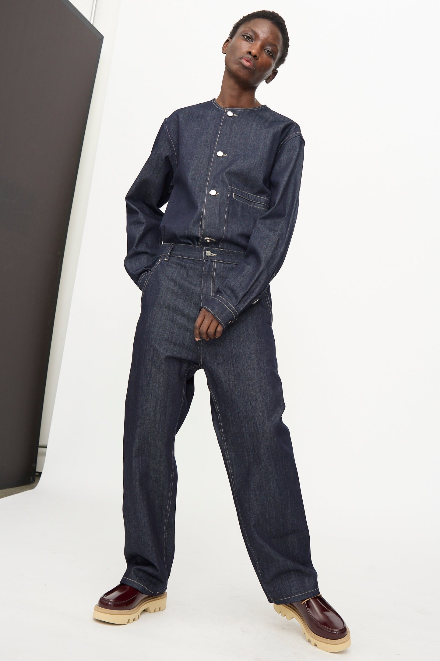 A person stands in a studio wearing Sofie D'Hoore's Raw Denim Pasay Pant in Midnight with a relaxed fit jacket, matched with maroon shoes featuring thick beige soles.