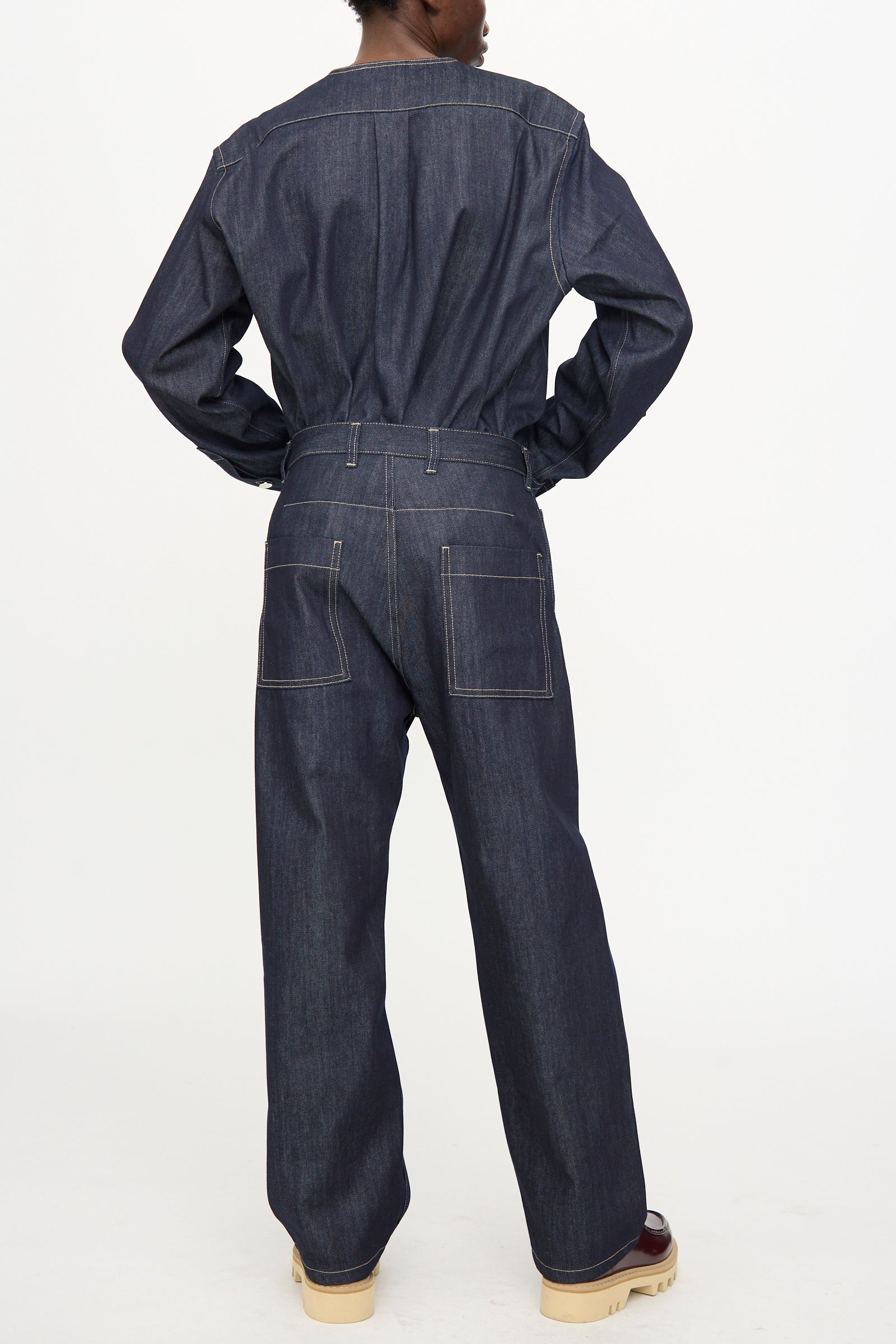 Person stands with back to camera, wearing Sofie D'Hoore's Raw Denim Pasay Pant in Midnight—a relaxed fit, straight leg dark denim—paired with light-colored shoes against a plain white background.
