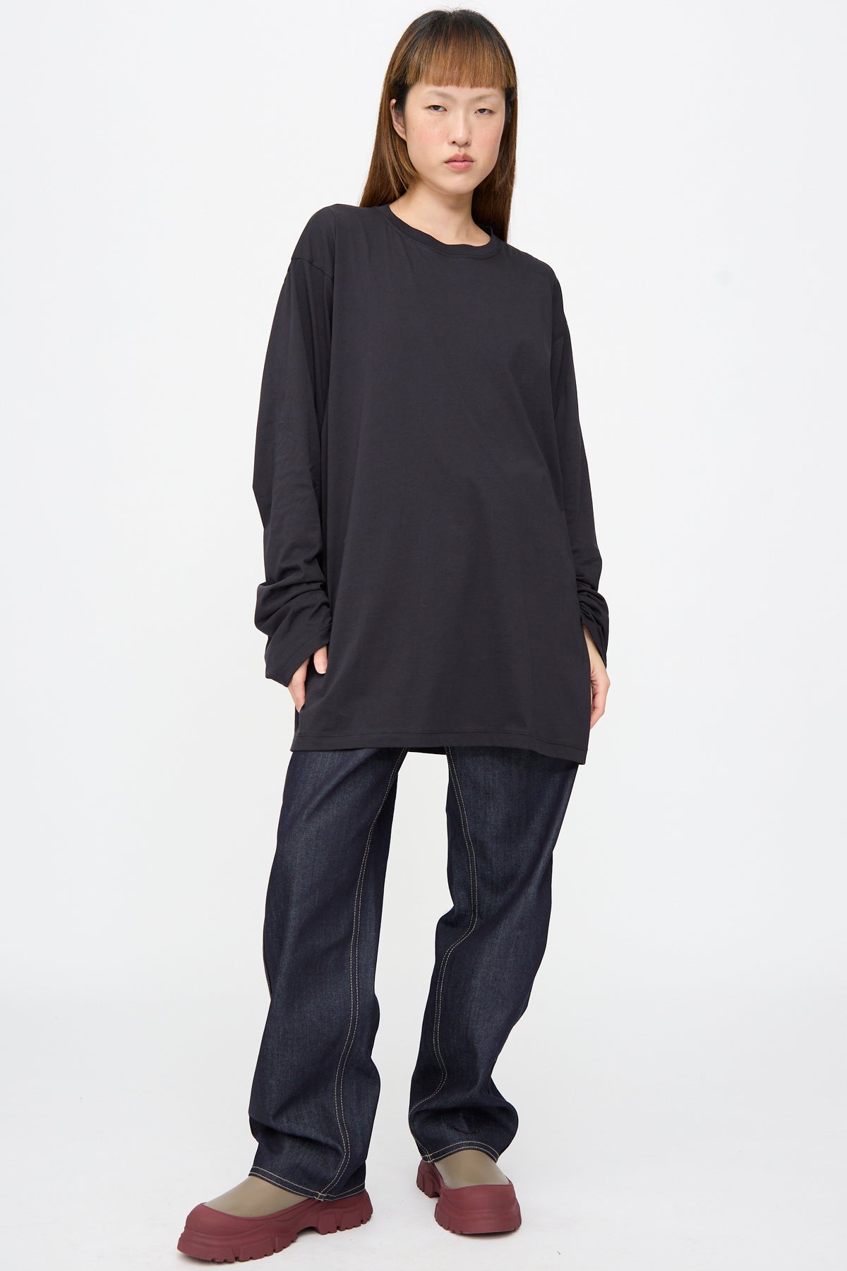 A person wearing an oversized black long-sleeve T-shirt, Sofie D’Hoore Raw Denim Phill Pant in Midnight with contrast stitching, and chunky olive-green and burgundy platform shoes.