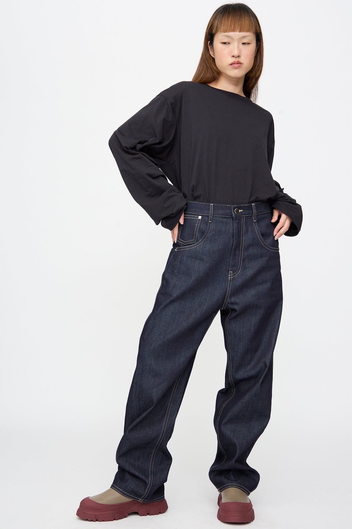 A person wearing a long-sleeved black shirt and Sofie D’Hoore Raw Denim Phill Pant in Midnight with contrast stitching, paired with olive-green boots with thick red soles.