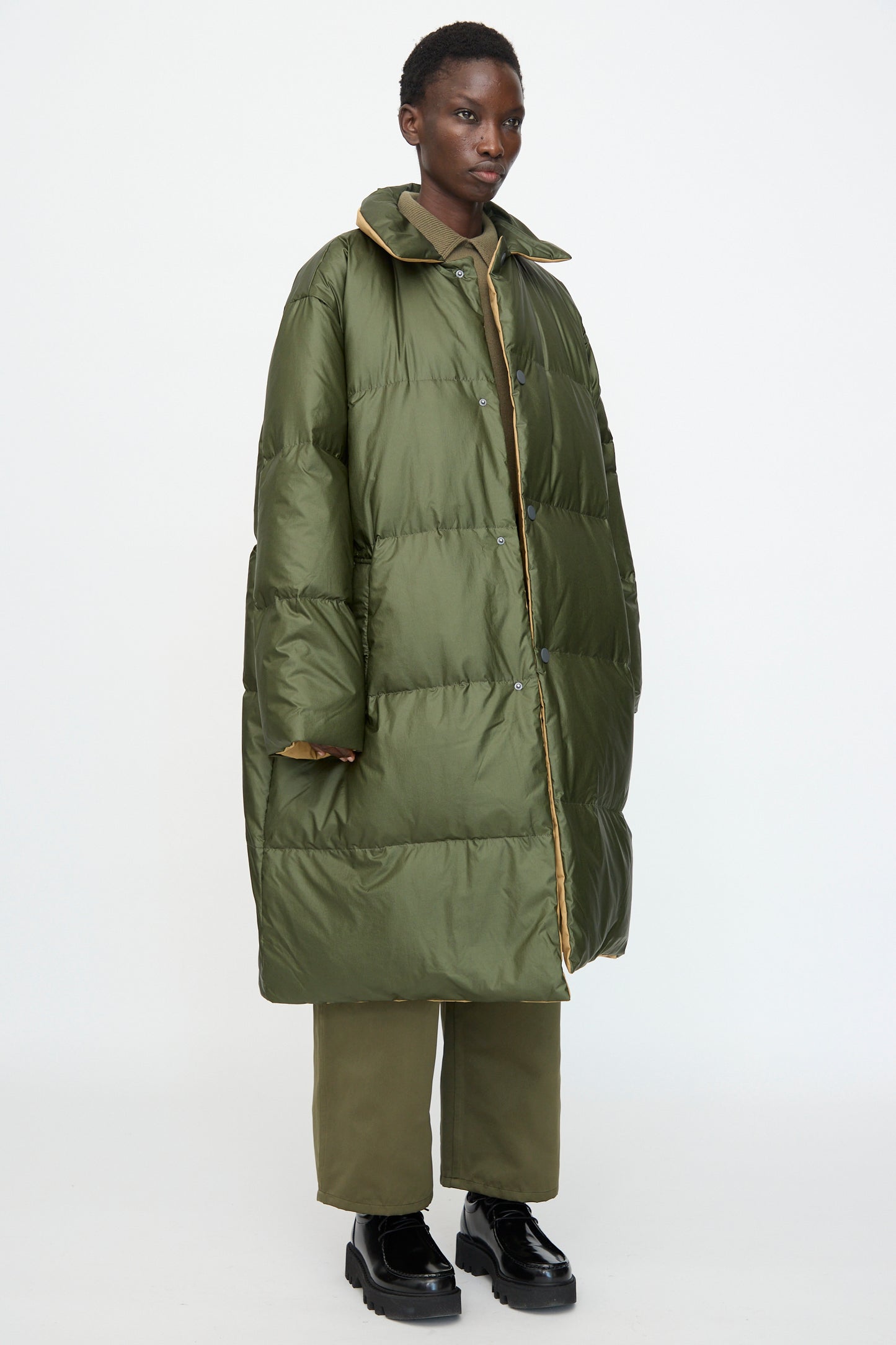 A person wears the Sofie D'Hoore Reversible Otis Down Coat in Mushroom and Khaki with green pants and black shoes, standing against a plain white background.