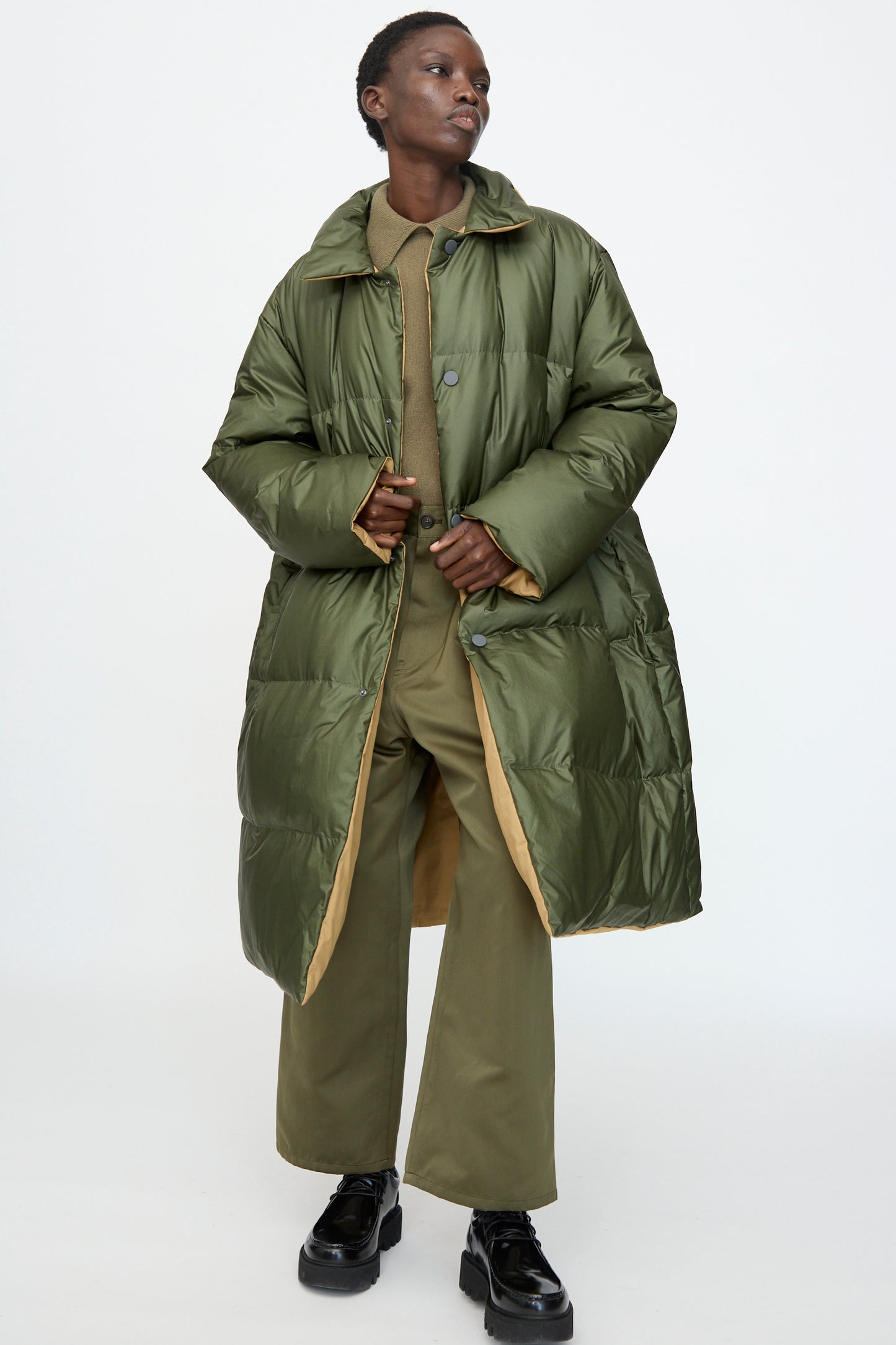A model wears the Sofie D'Hoore Reversible Otis Down Coat in Mushroom and Khaki over a green outfit with black platform shoes, posed against a plain white background.