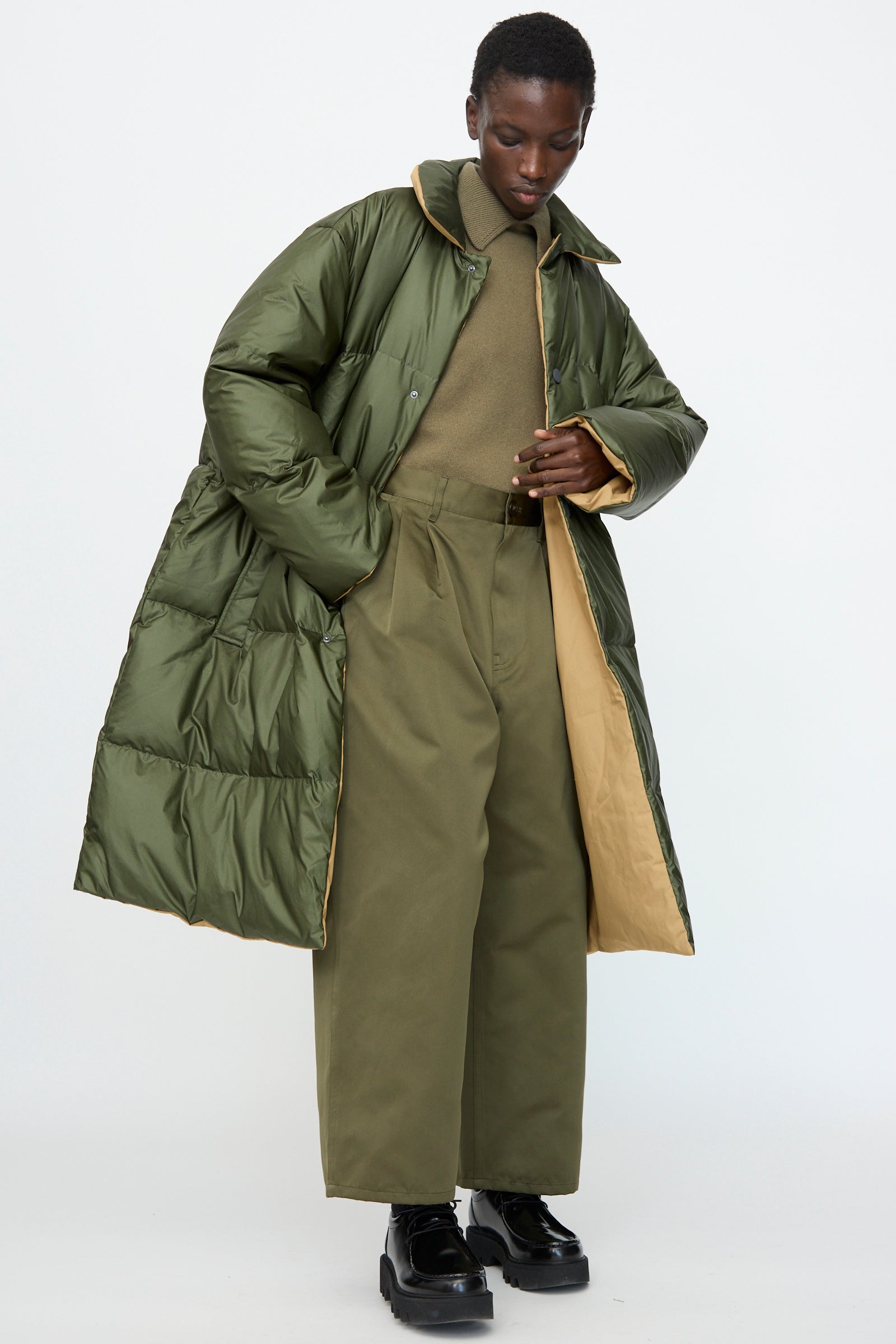 A person wearing the Sofie D'Hoore Reversible Otis Down Coat in Mushroom and Khaki stands against a plain light background, paired with a matching turtleneck, wide-leg trousers, and black platform shoes.