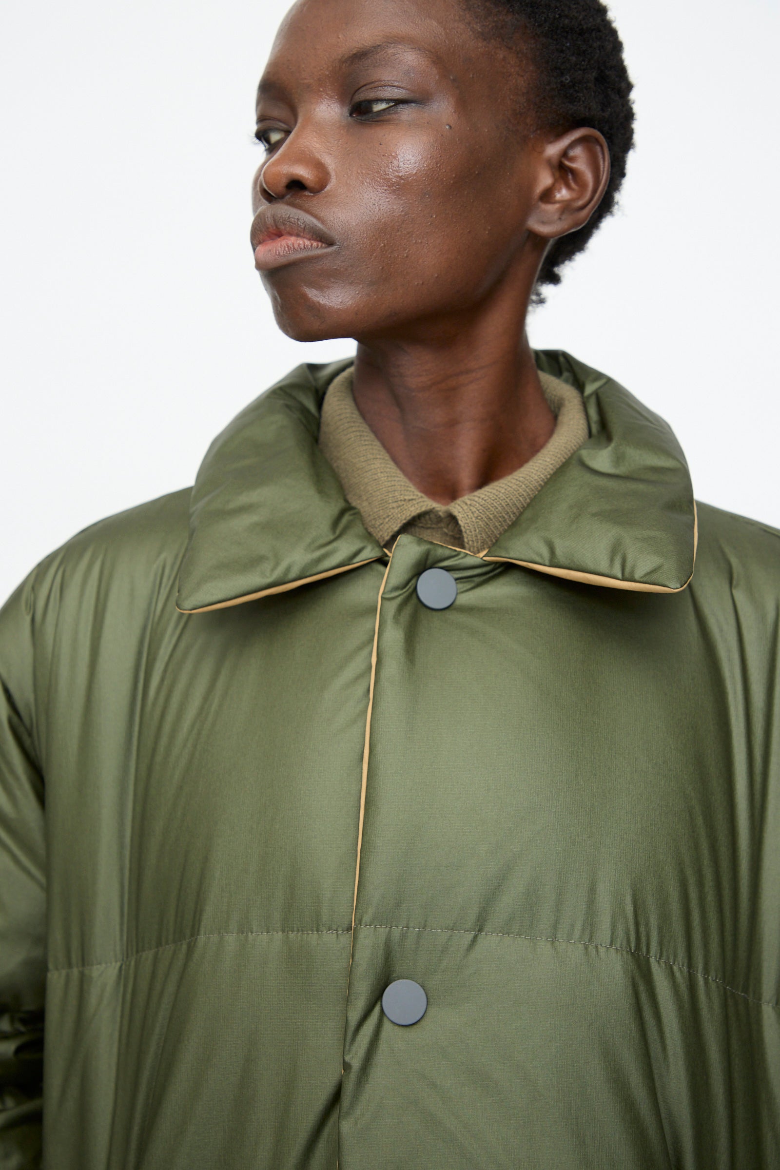 A person wearing the Sofie D'Hoore Reversible Otis Down Coat in Mushroom and Khaki over a light brown shirt, looking to the side against a plain background.
