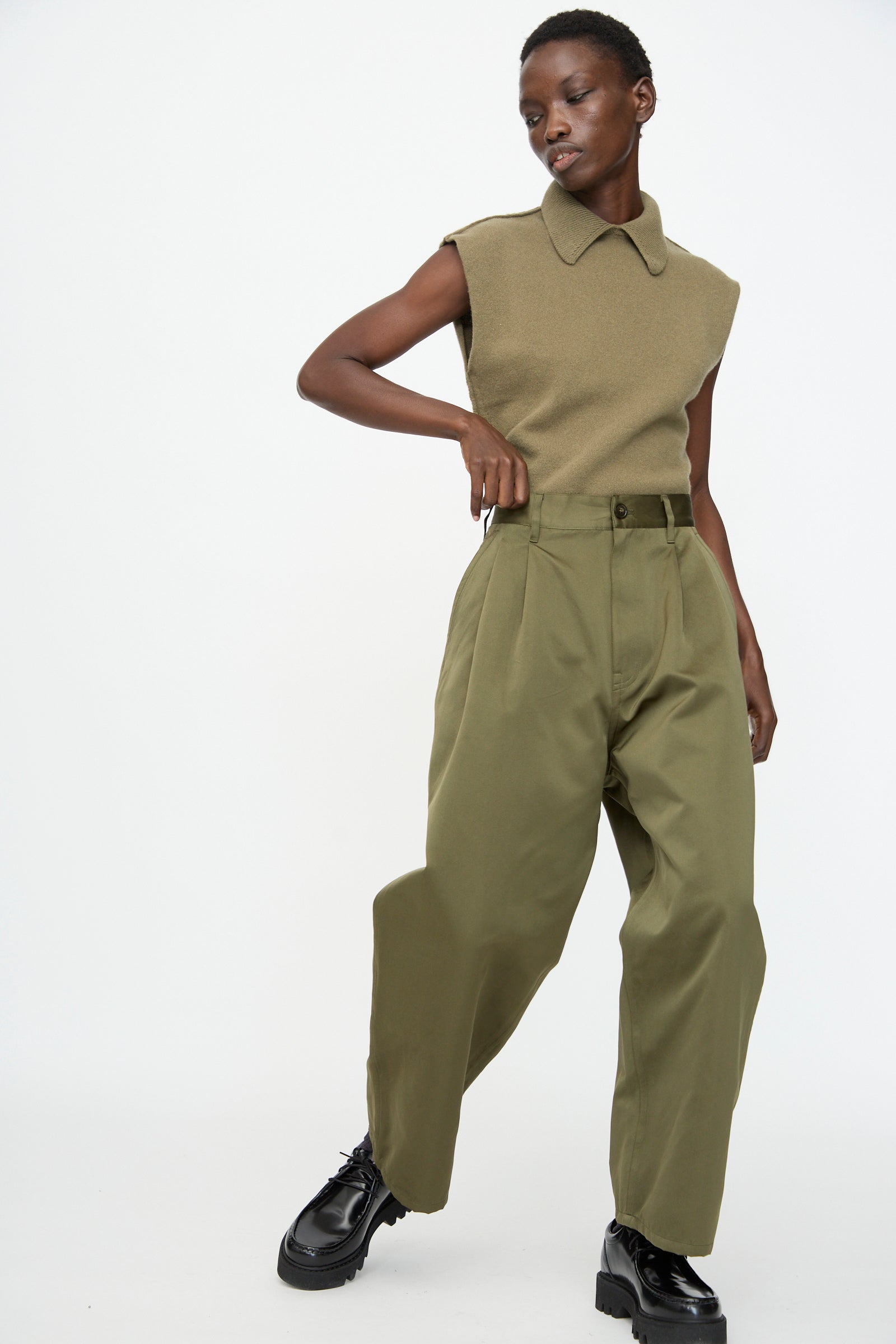 A person wearing a sleeveless collared olive top, Sofie D'Hoore Satin Polke Pant in Laurel, and black chunky shoes stands against a plain white background.