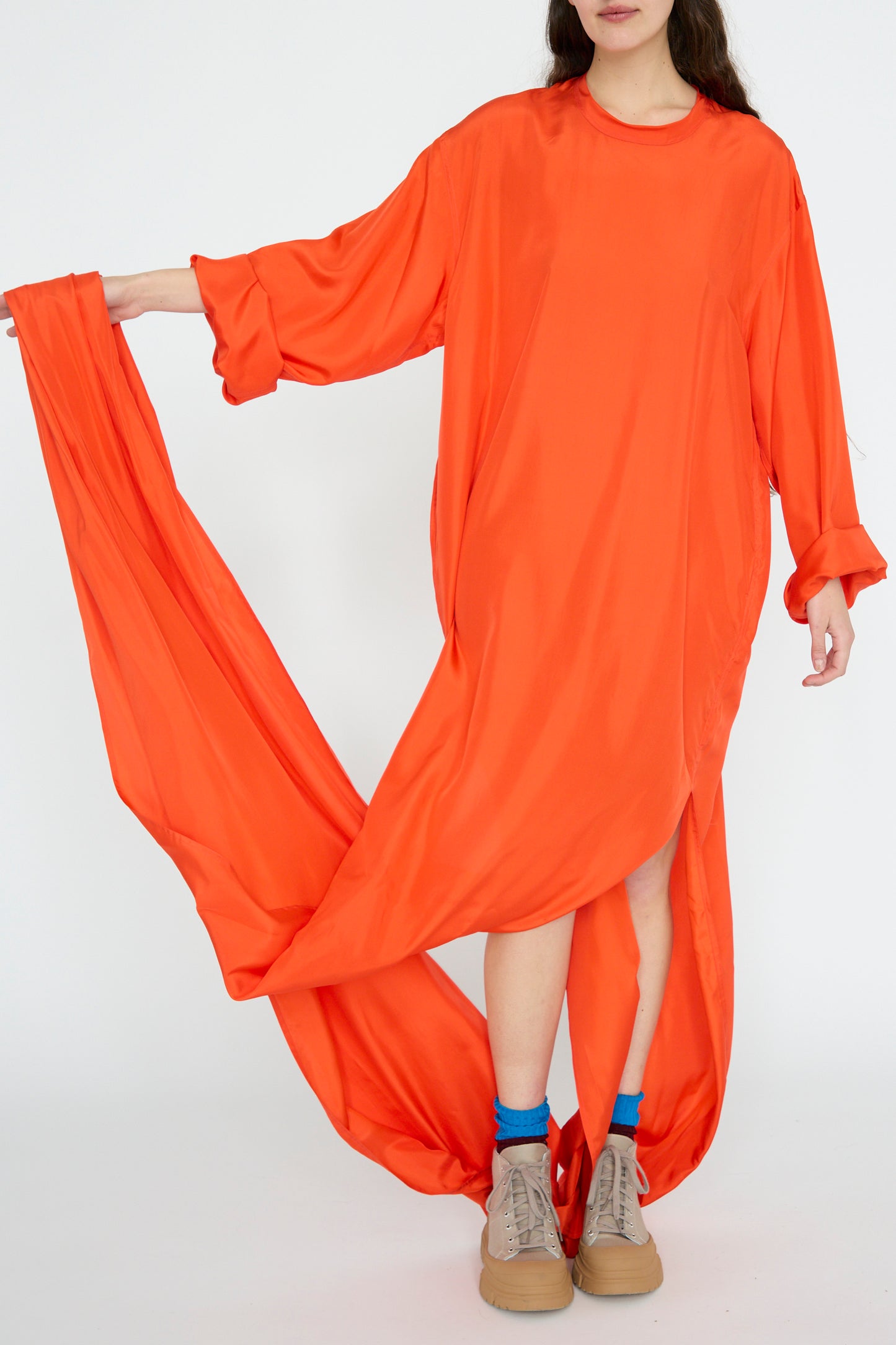 A person stands against a plain background wearing the Sofie D'Hoore Silk Habotai Dolphin Dress in Poppy, known for its long, flowing design with an oversize fit. The look is completed with tan shoes and blue socks.