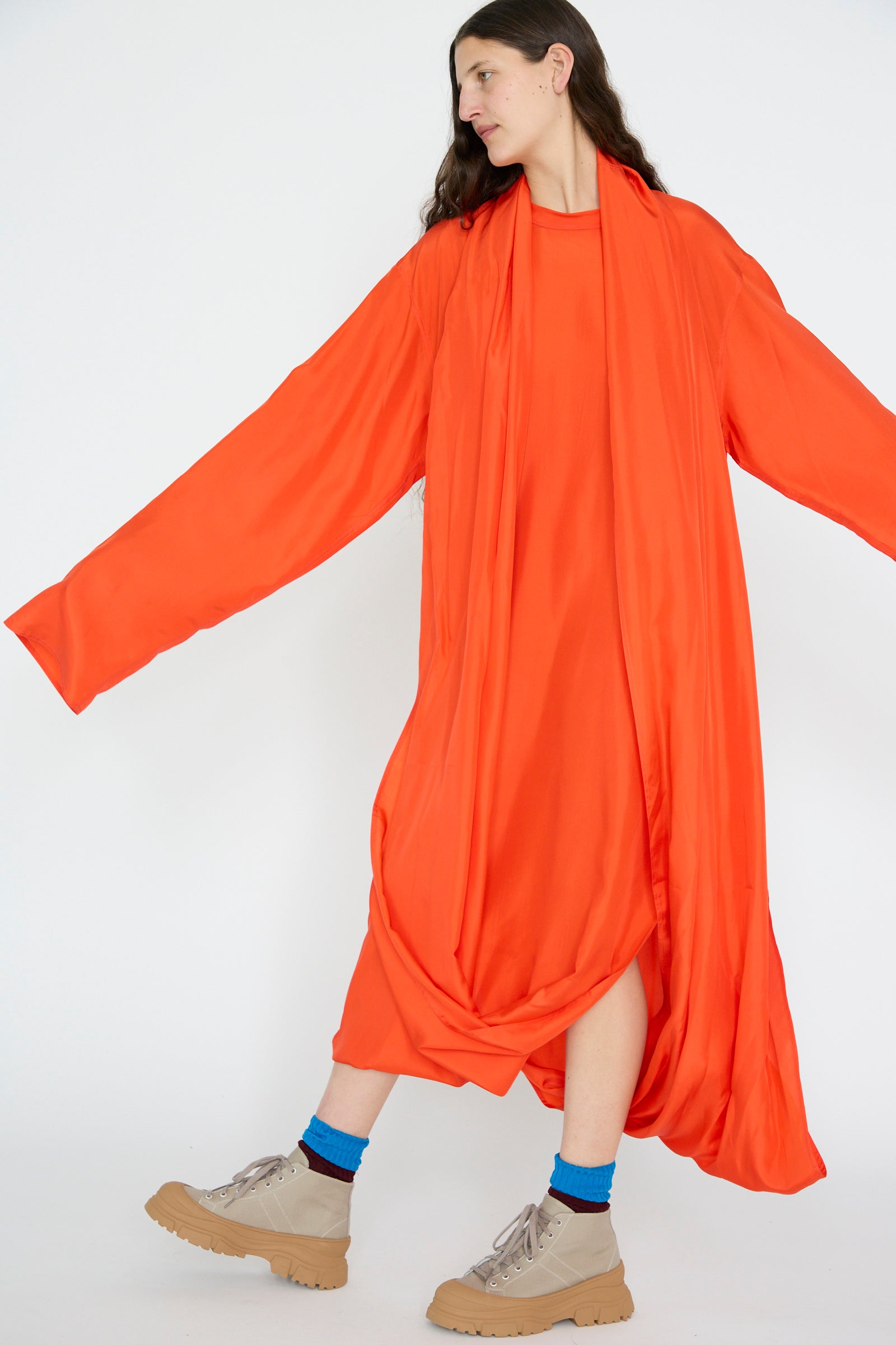 A person stands against a white background wearing the Silk Habotai Dolphin Dress in Poppy by Sofie D'Hoore, an oversize, long-sleeve flowing orange dress, paired with tan boots.