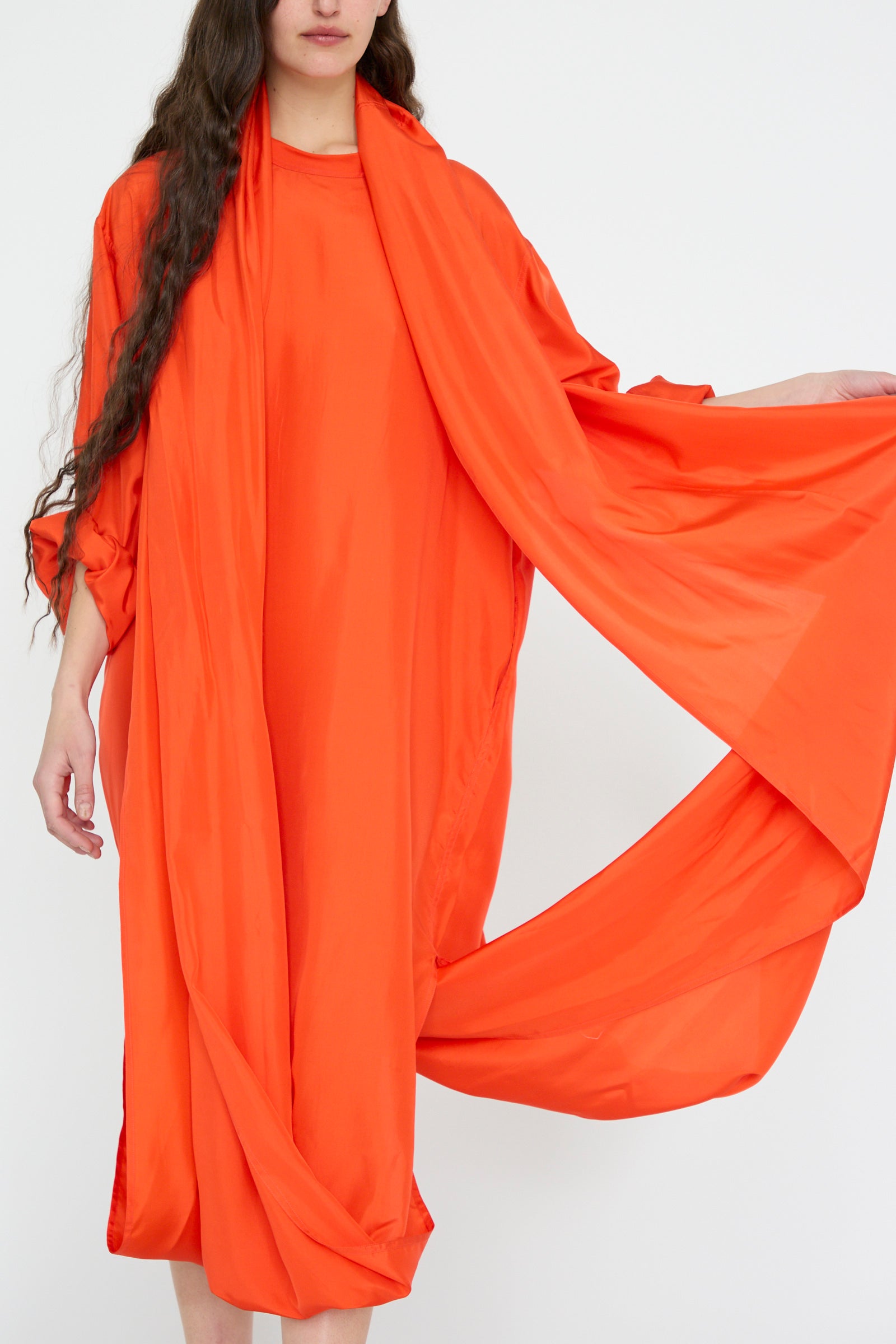 A person models the Silk Habotai Dolphin Dress in Poppy by Sofie D'Hoore, featuring an oversize fit and long draped fabric, against a plain white backdrop.