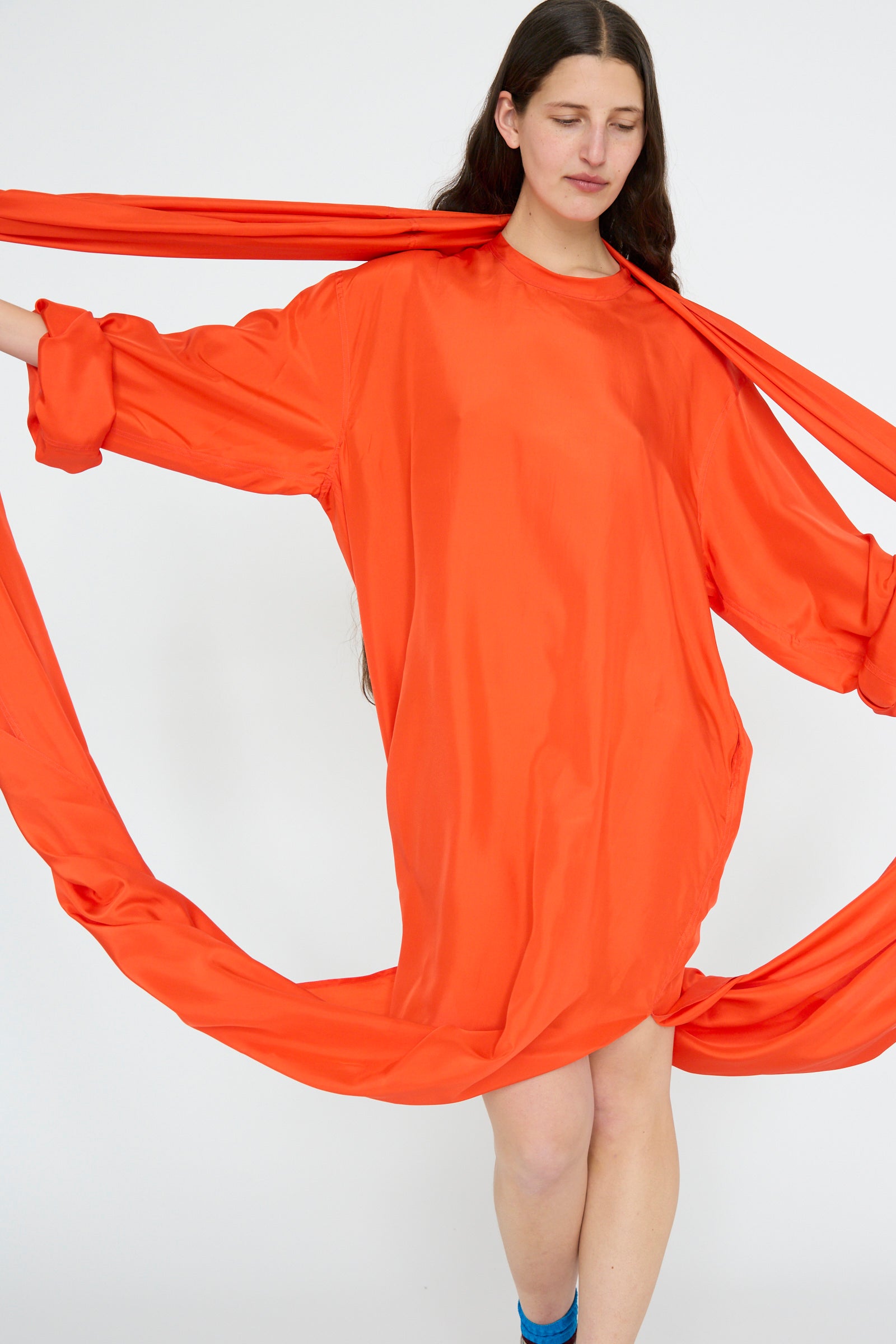 A person poses against a plain background wearing the Silk Habotai Dolphin Dress in Poppy by Sofie D'Hoore, showcasing long sleeves, a loose fit, and  draped silk Habotai fabric around the arms.