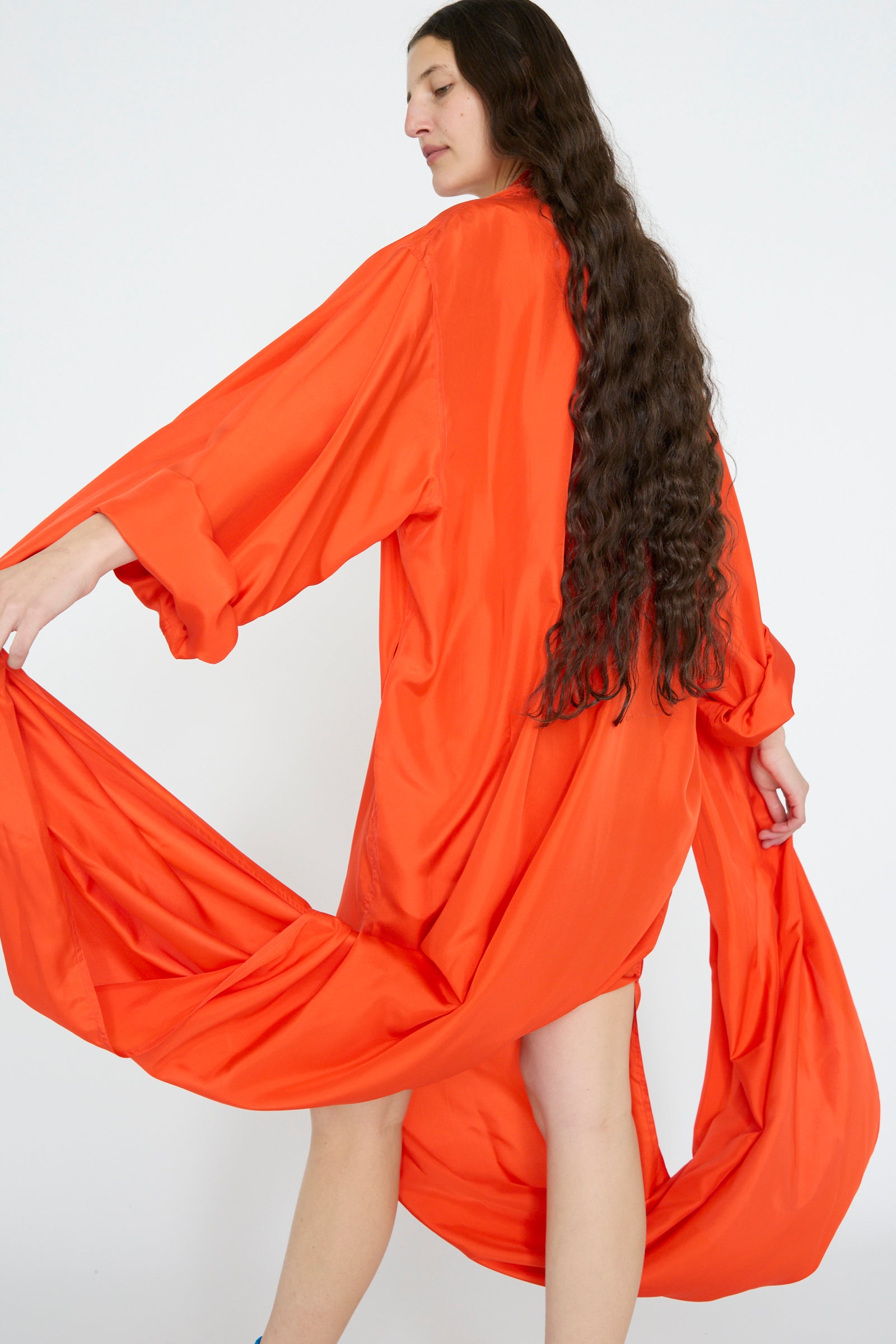 Against a white background, a person with long hair stands facing away in an oversized Silk Habotai Dolphin Dress in Poppy by Sofie D'Hoore. 