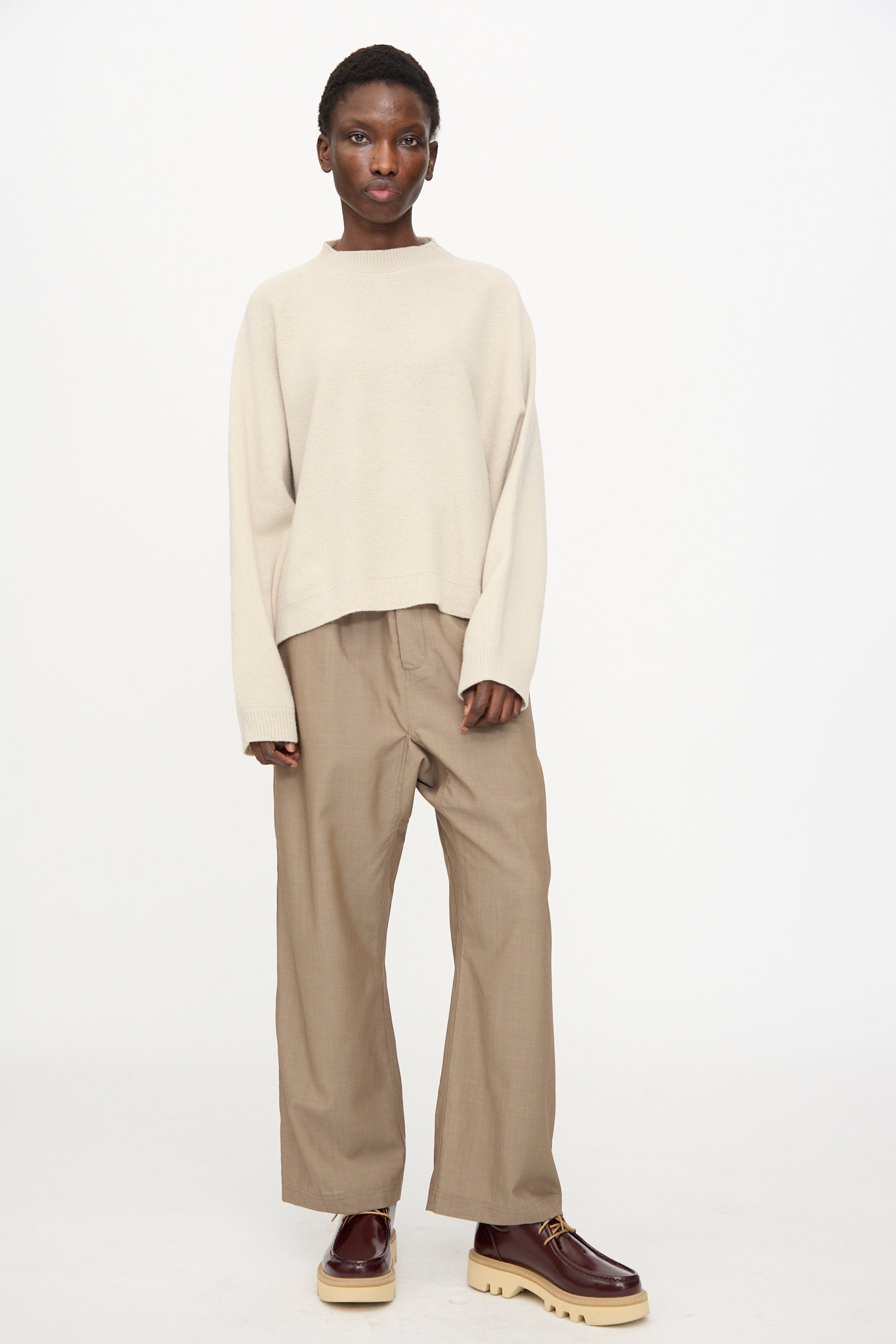 A person stands against a white background wearing the Sofie D'Hoore Superfine Wool Maui Sweater in Natural, paired with tan wide-leg pants and burgundy platform shoes.
