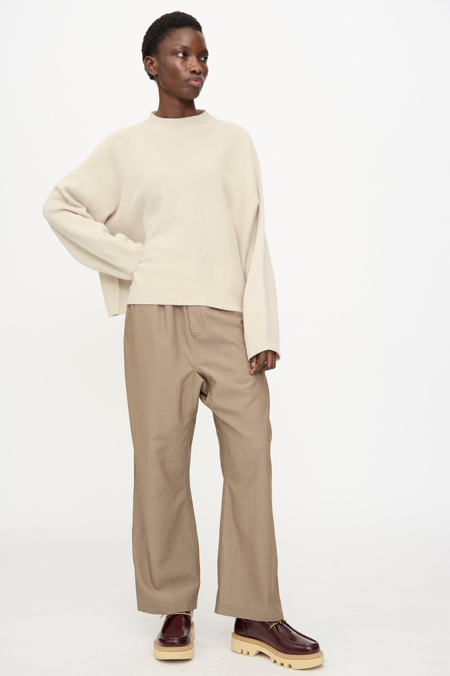 A person stands against a white background, wearing the Sofie D'Hoore Superfine Wool Maui Sweater in Natural, paired with tan wide-leg pants and brown platform shoes.