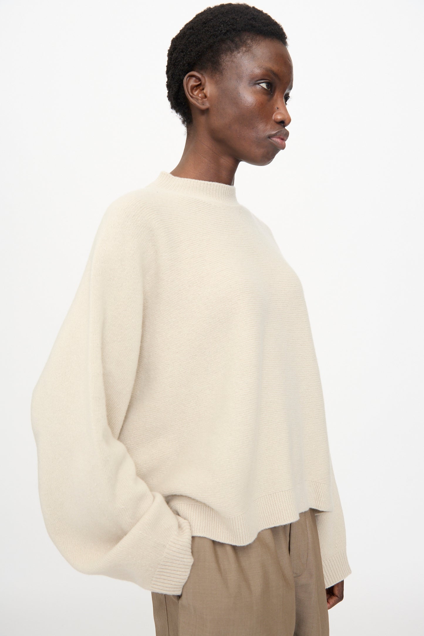 A person wears the Sofie D'Hoore Superfine Wool Maui Sweater in Natural, a relaxed fit, light beige oversized pullover, paired with tan pants and stands against a plain white background, facing slightly to the side.