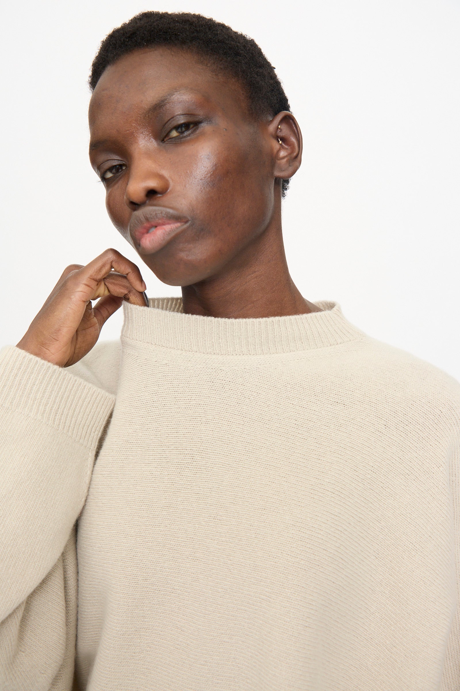 A person wears the Sofie D'Hoore Superfine Wool Maui Sweater in Natural, holding the collar with one hand and gazing at the camera with a neutral expression against a plain background.