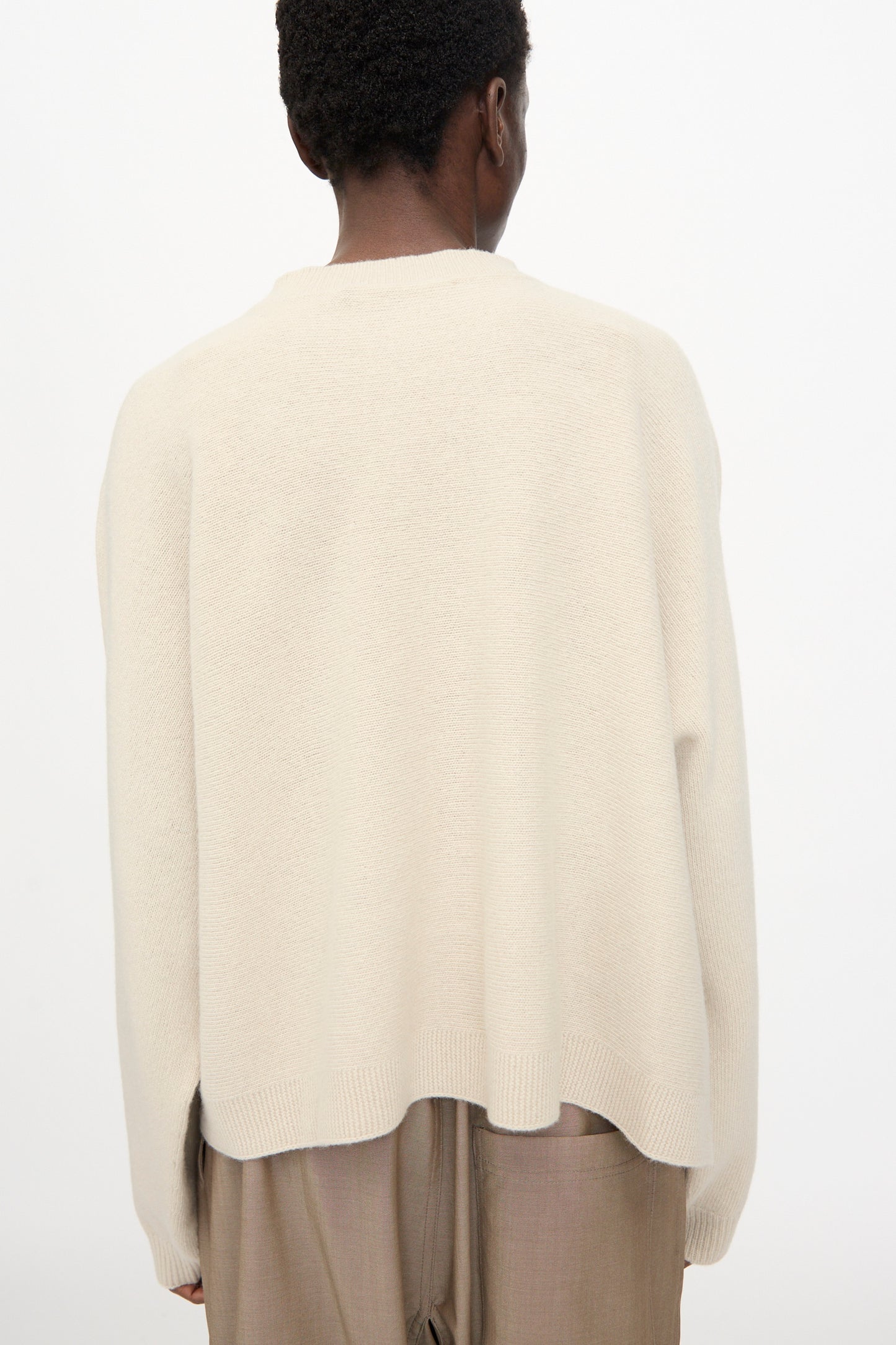 Person stands with their back to the camera, wearing the Sofie D'Hoore Superfine Wool Maui Sweater in Natural—an oversized, relaxed fit pullover—paired with light brown pants against a plain, light background.
