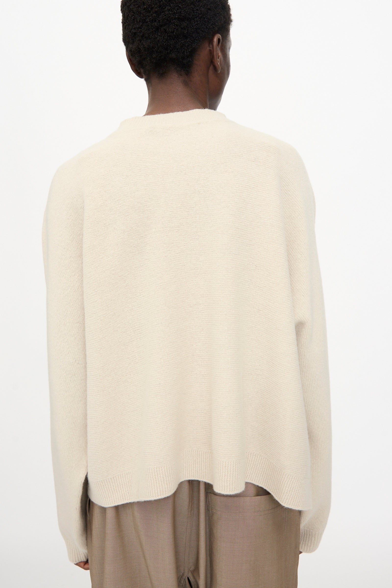 Person stands with their back to the camera, wearing the Sofie D'Hoore Superfine Wool Maui Sweater in Natural—an oversized, relaxed fit pullover—paired with light brown pants against a plain, light background.