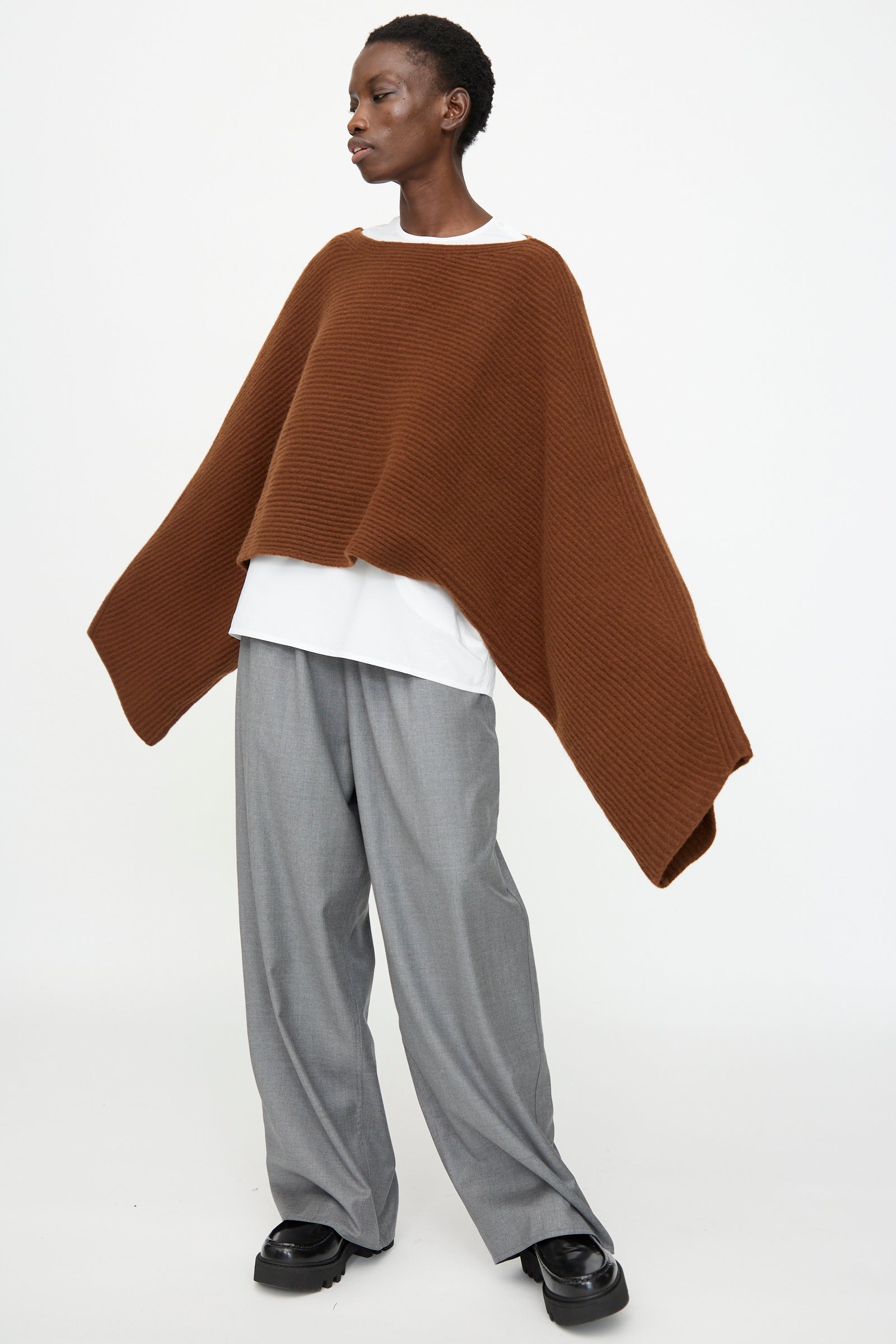 A person wears the Sofie D'Hoore Superfine Wool Murakami Oversized Sweater in Chestnut over a white shirt, gray wide-leg trousers, and black shoes while standing against a plain white background.