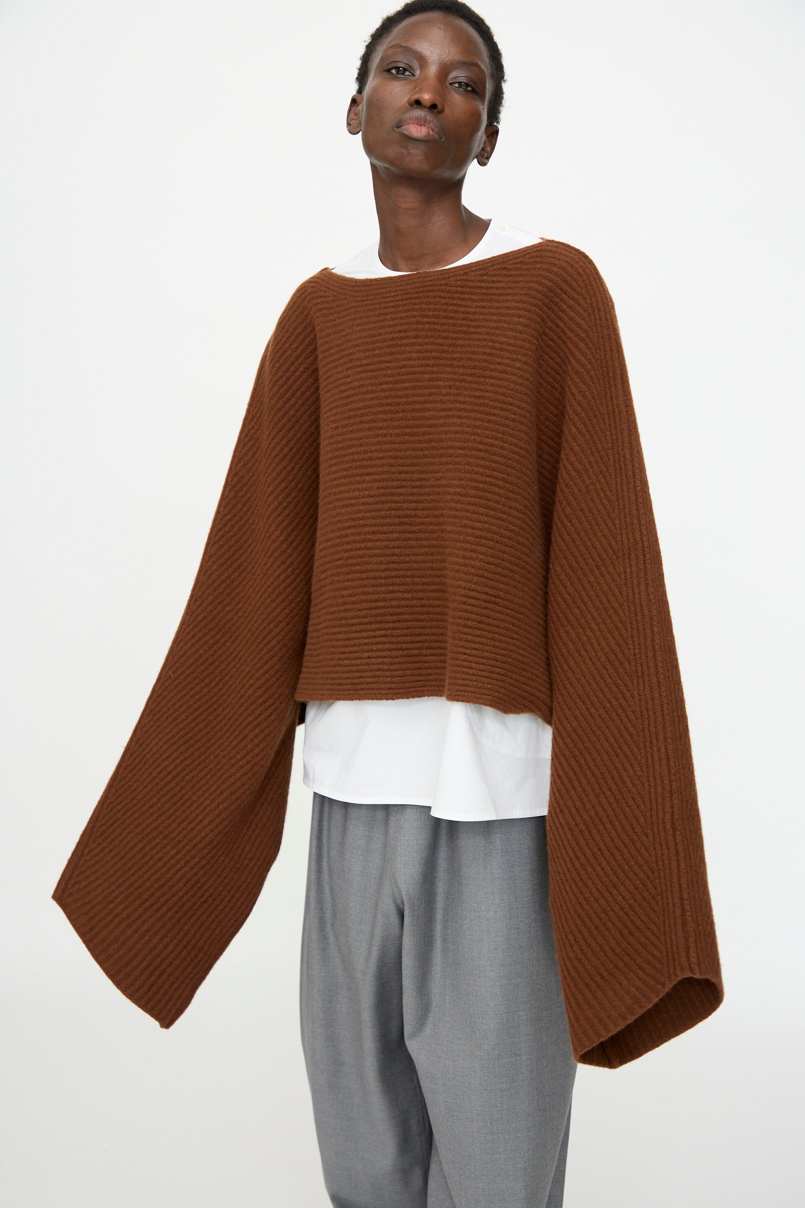 Model wears the Sofie D'Hoore Superfine Wool Murakami Oversized Sweater in Chestnut, styled with a white shirt and gray pants, standing against a plain white background.