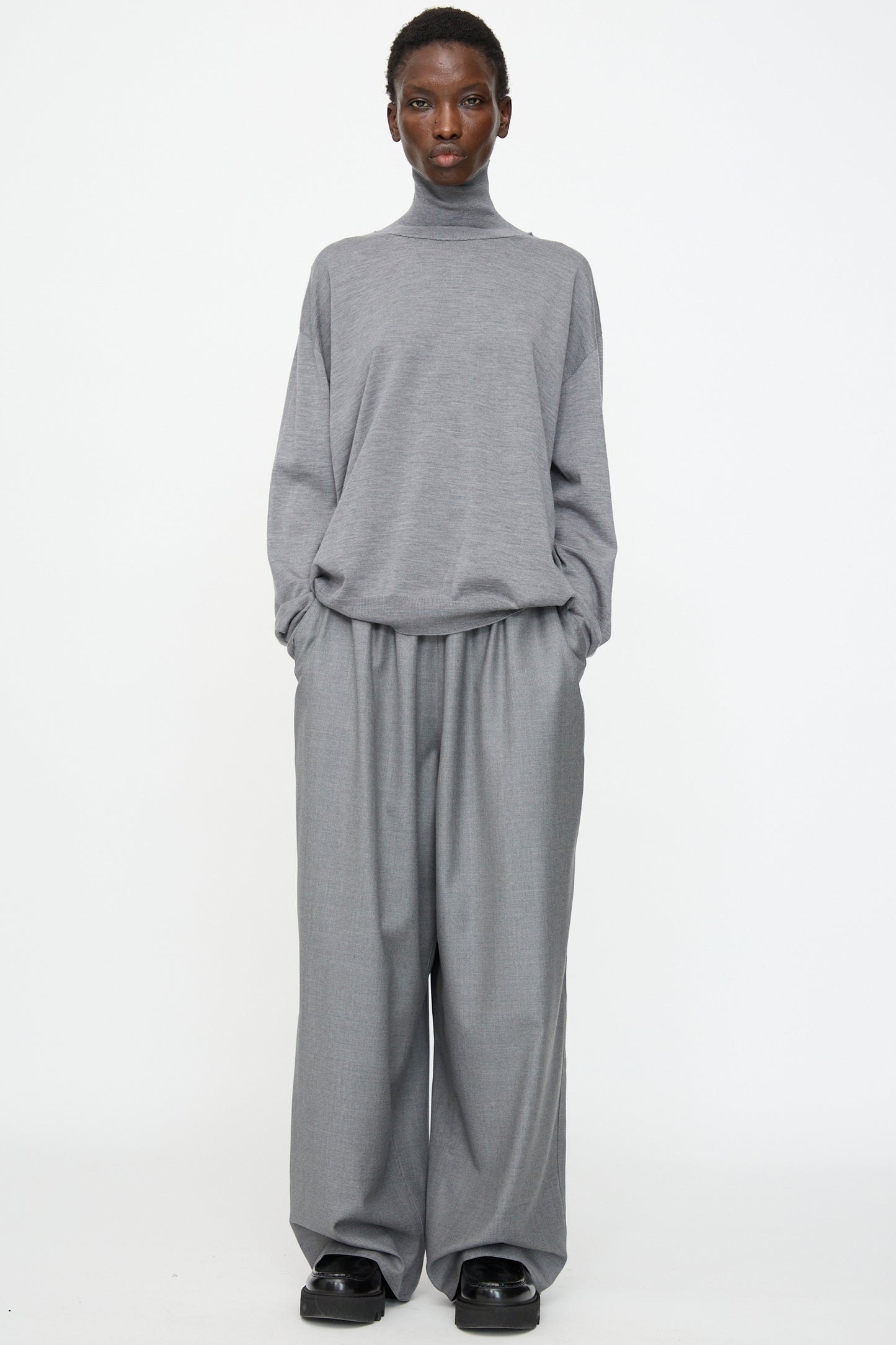 A person faces forward against a plain white background, wearing Sofie D'Hoore's Superfine Wool Pasch Pants in Mid Grey Melange with an elastic waistband, paired with a loose-fitting grey turtleneck sweater and black shoes.
