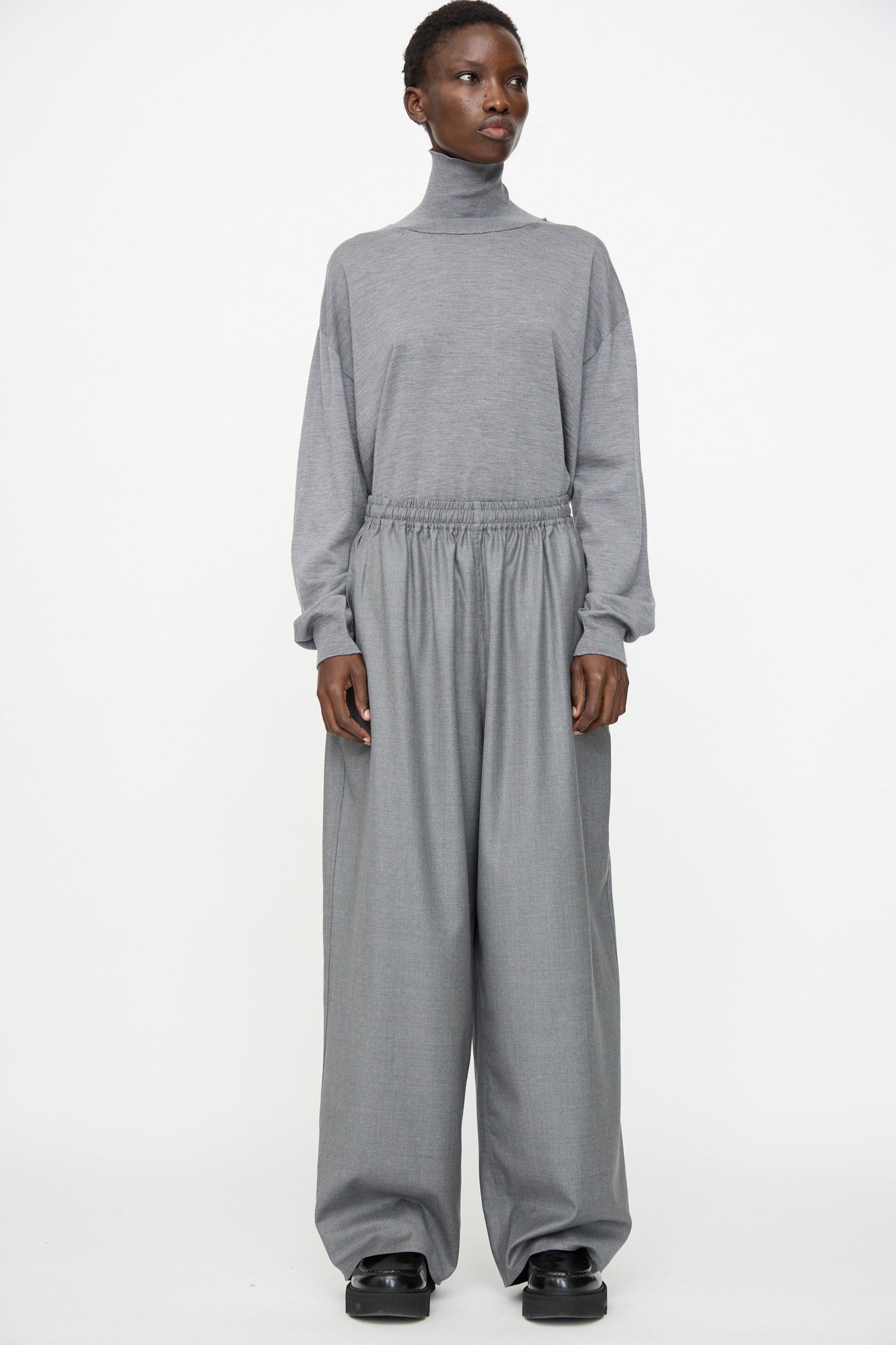A person stands against a plain background, wearing a grey turtleneck top and Sofie D'Hoore Superfine Wool Pasch Pants in Mid Grey Melange, styled with black shoes.