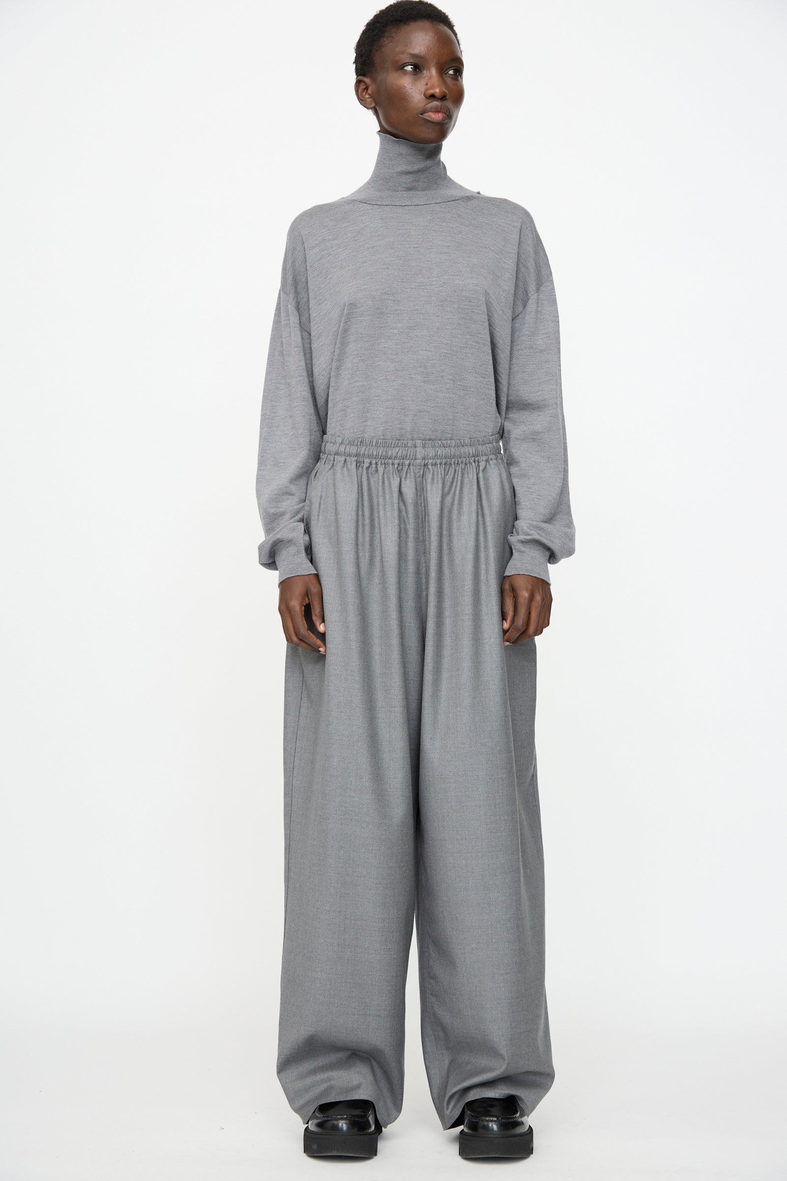A person stands against a plain background, wearing a grey turtleneck top and Sofie D'Hoore Superfine Wool Pasch Pants in Mid Grey Melange, styled with black shoes.