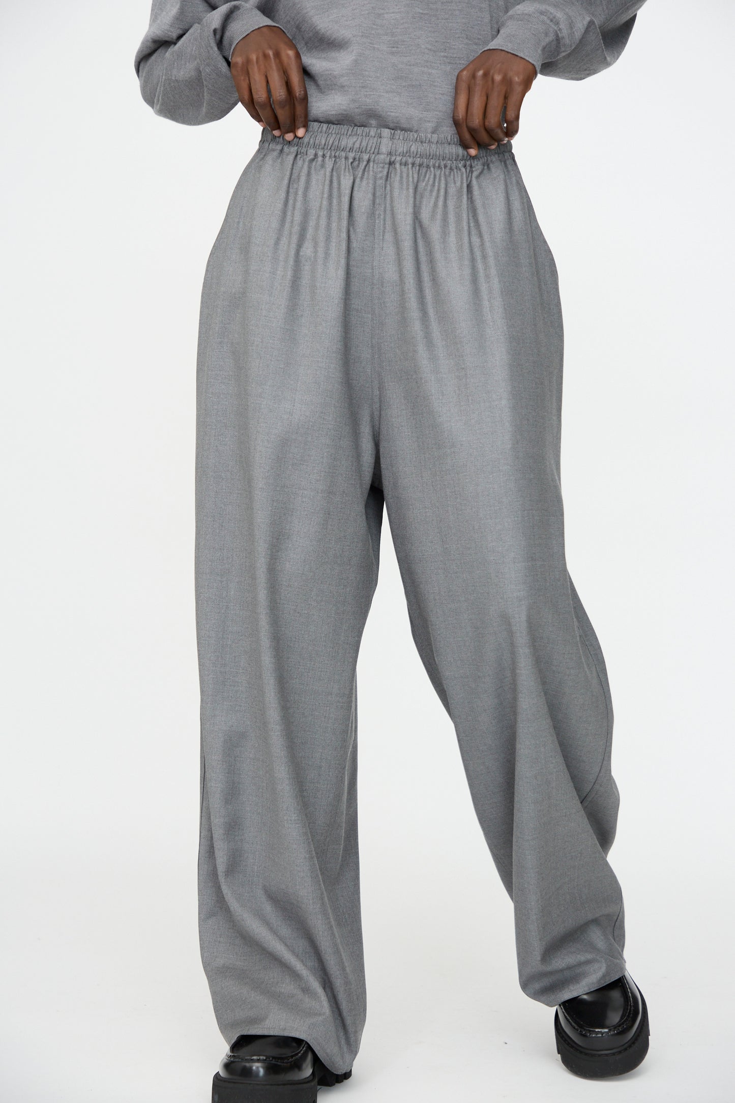 A person wears Sofie D'Hoore Superfine Wool Pasch Pants in Mid Grey Melange, featuring an elastic waistband, paired with black shoes, standing against a plain white background.