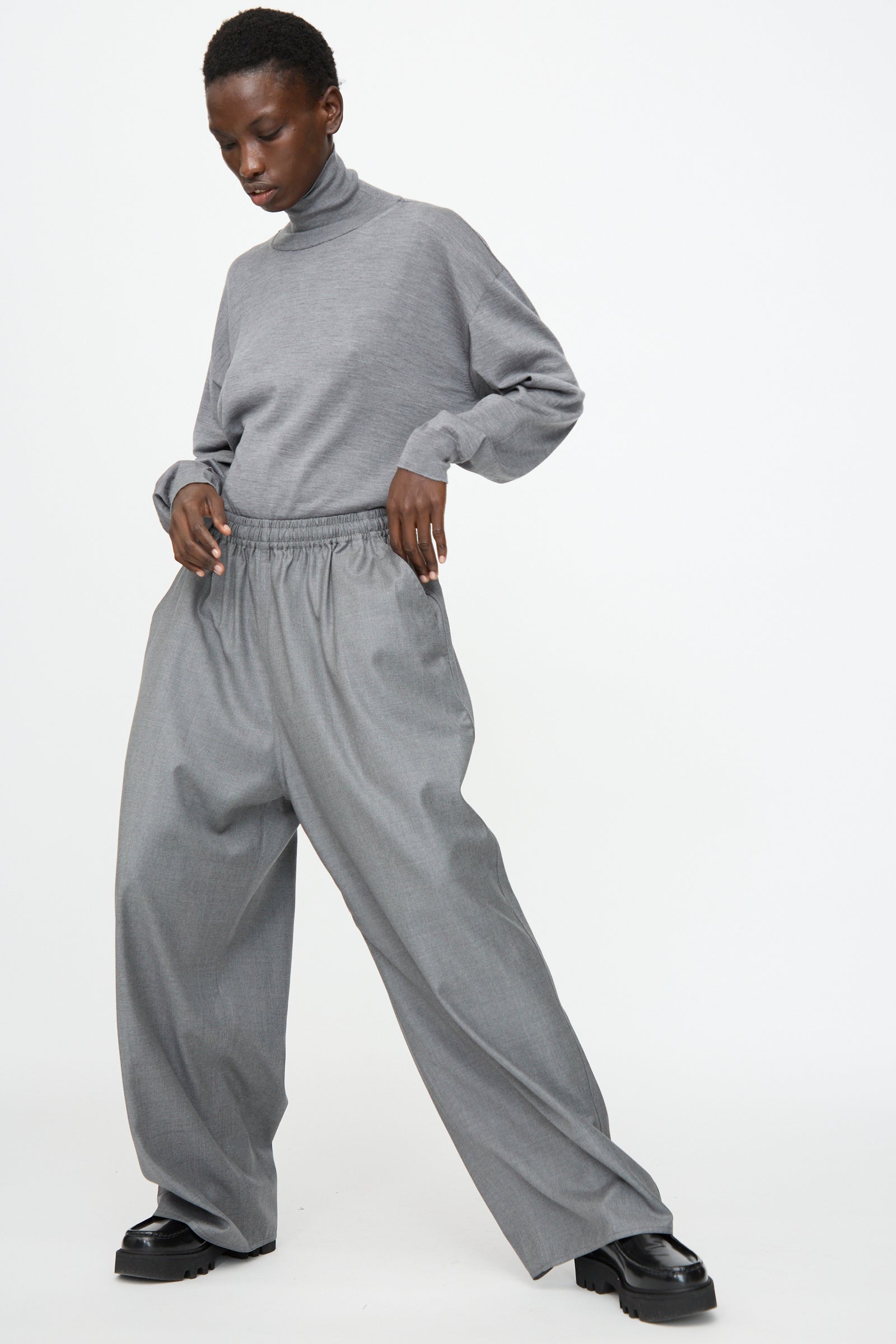 A person wears the Sofie D’Hoore Superfine Wool Pasch Pants in Mid Grey Melange, styled with a loose grey turtleneck and black shoes, standing against a plain white background.