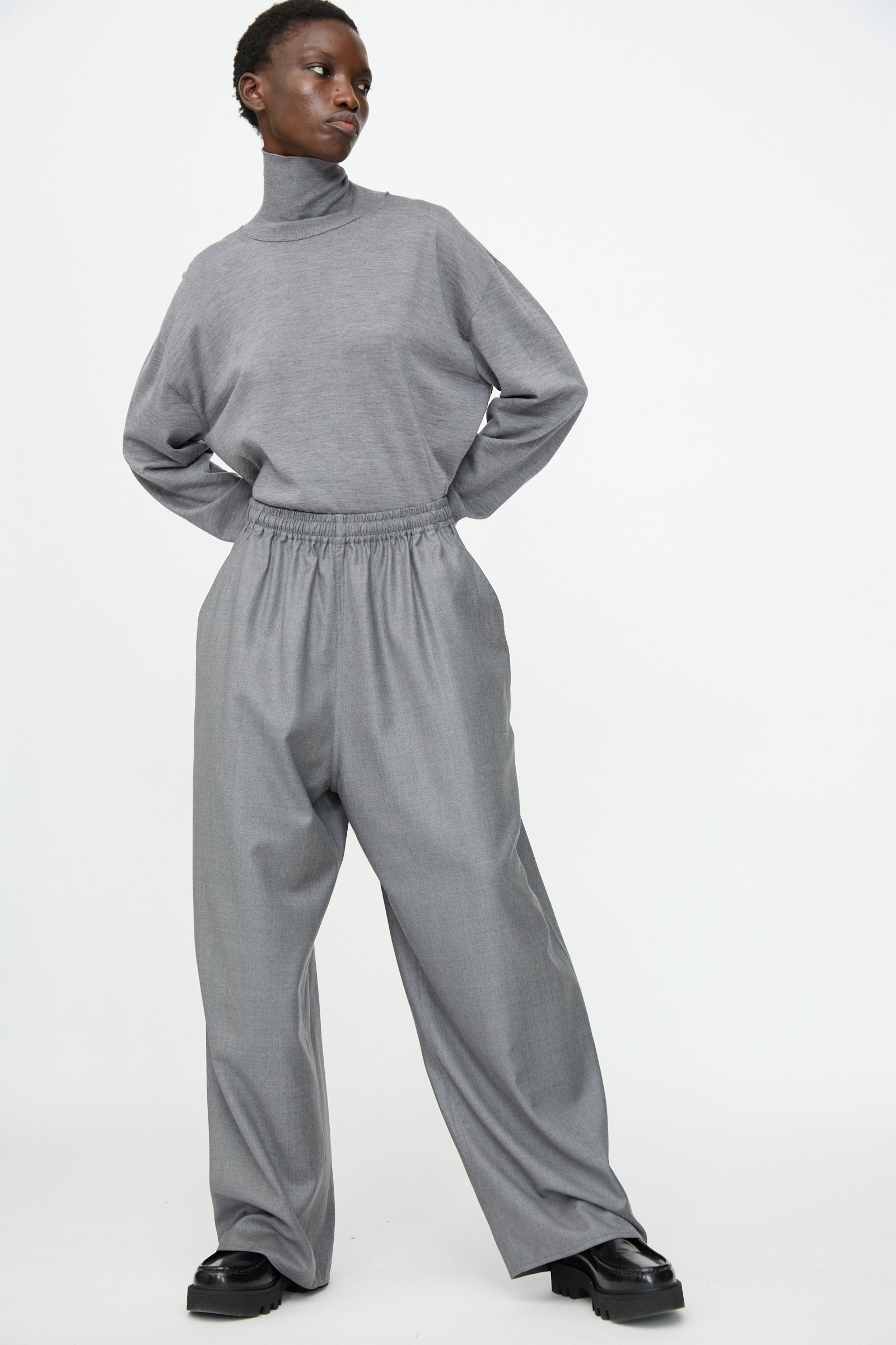 A person wears Sofie D'Hoore's Superfine Wool Pasch Pants in Mid Grey Melange with a loose, light-gray turtleneck and black shoes, standing against a plain white background.