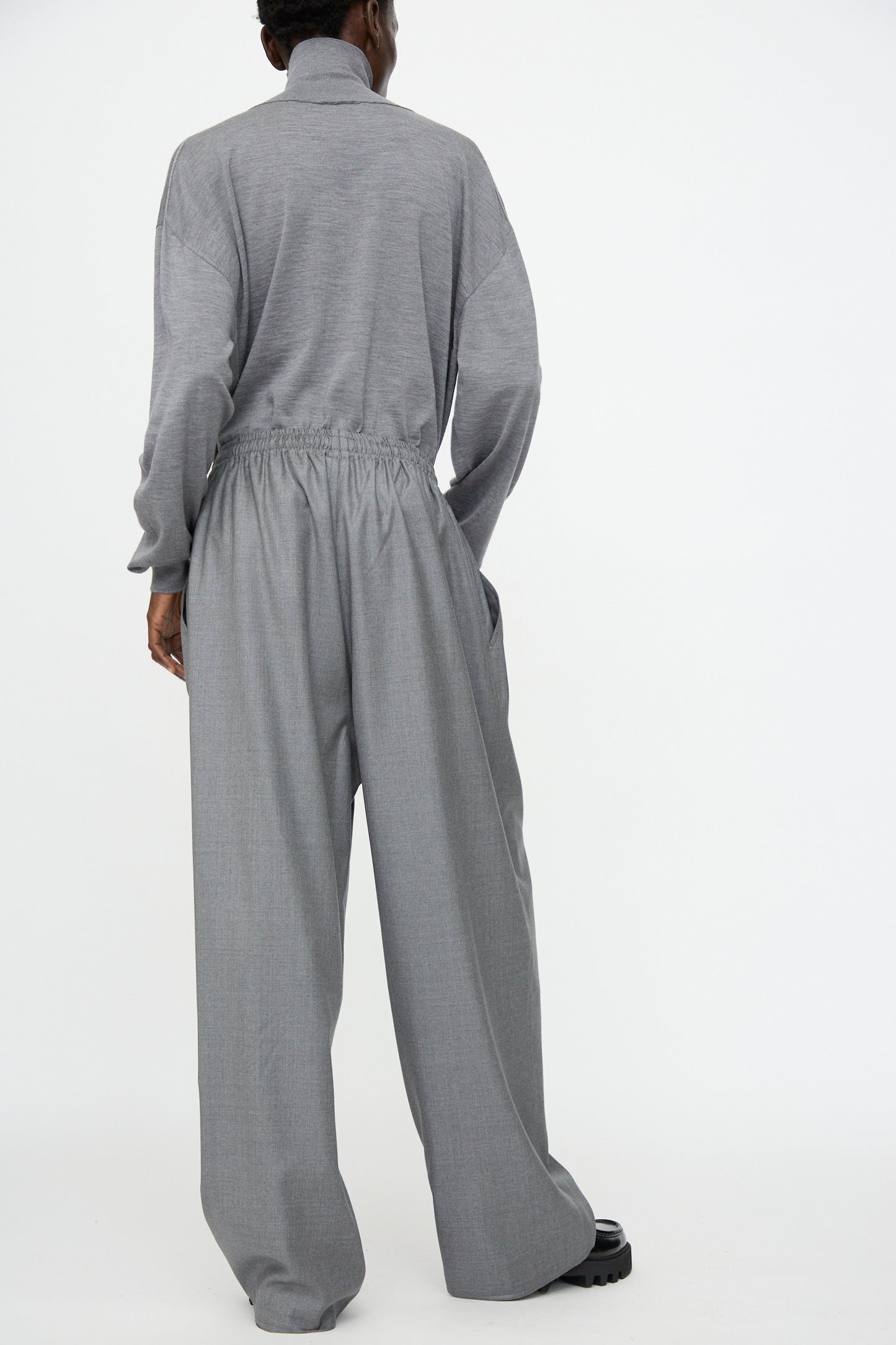 A person stands against a white background, shown from the back, wearing Sofie D'Hoore's Superfine Wool Pasch Pants in Mid Grey Melange with an elastic waistband and a loose-fitting, long-sleeved gray top.