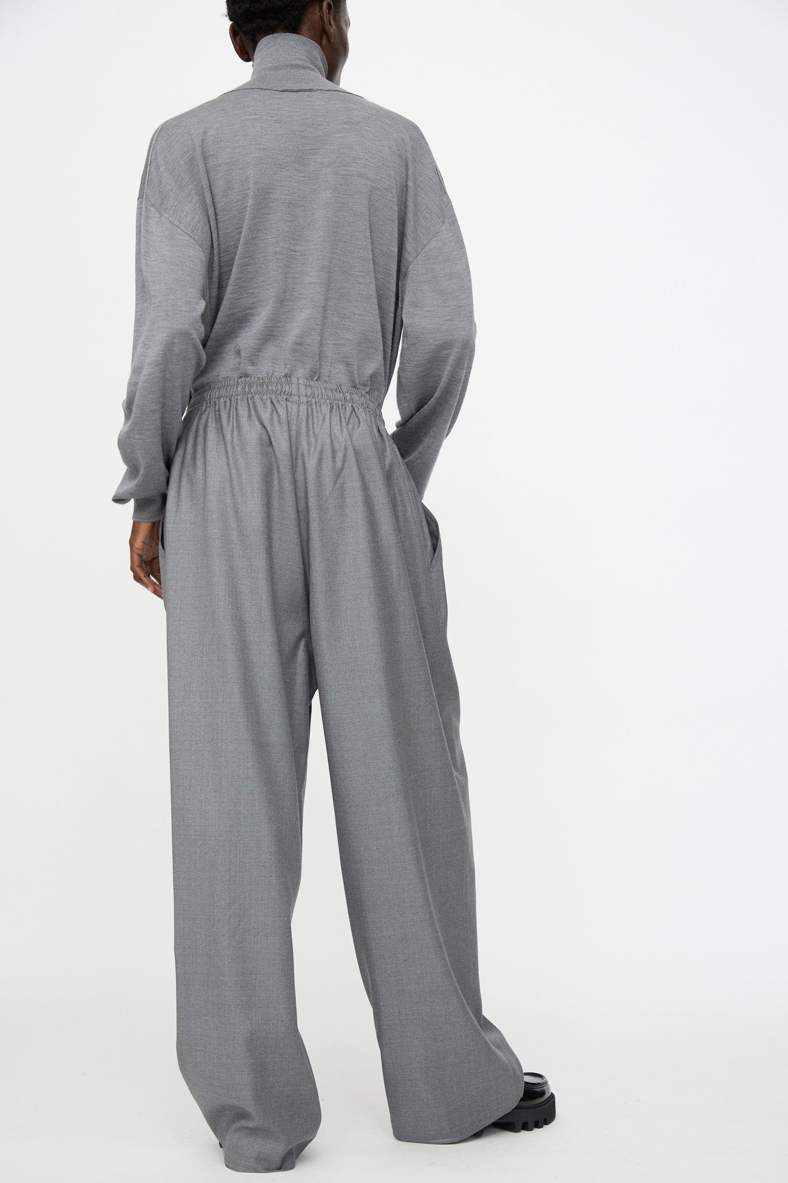 A person stands against a white background, shown from the back, wearing Sofie D'Hoore's Superfine Wool Pasch Pants in Mid Grey Melange with an elastic waistband and a loose-fitting, long-sleeved gray top.