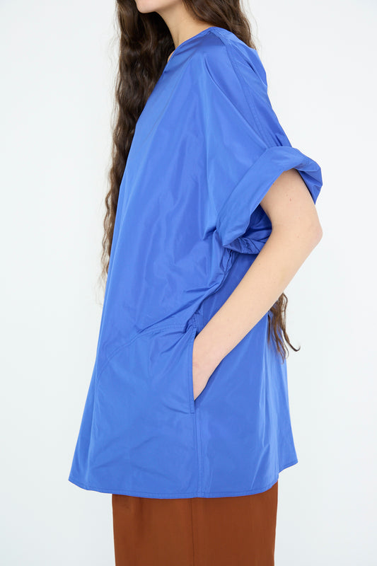 A person with long hair wears an oversize-fit Taffeta Buffalo Top in Sapphire by Sofie D'Hoore and brown pants, posing against a plain background with one hand in a pocket.